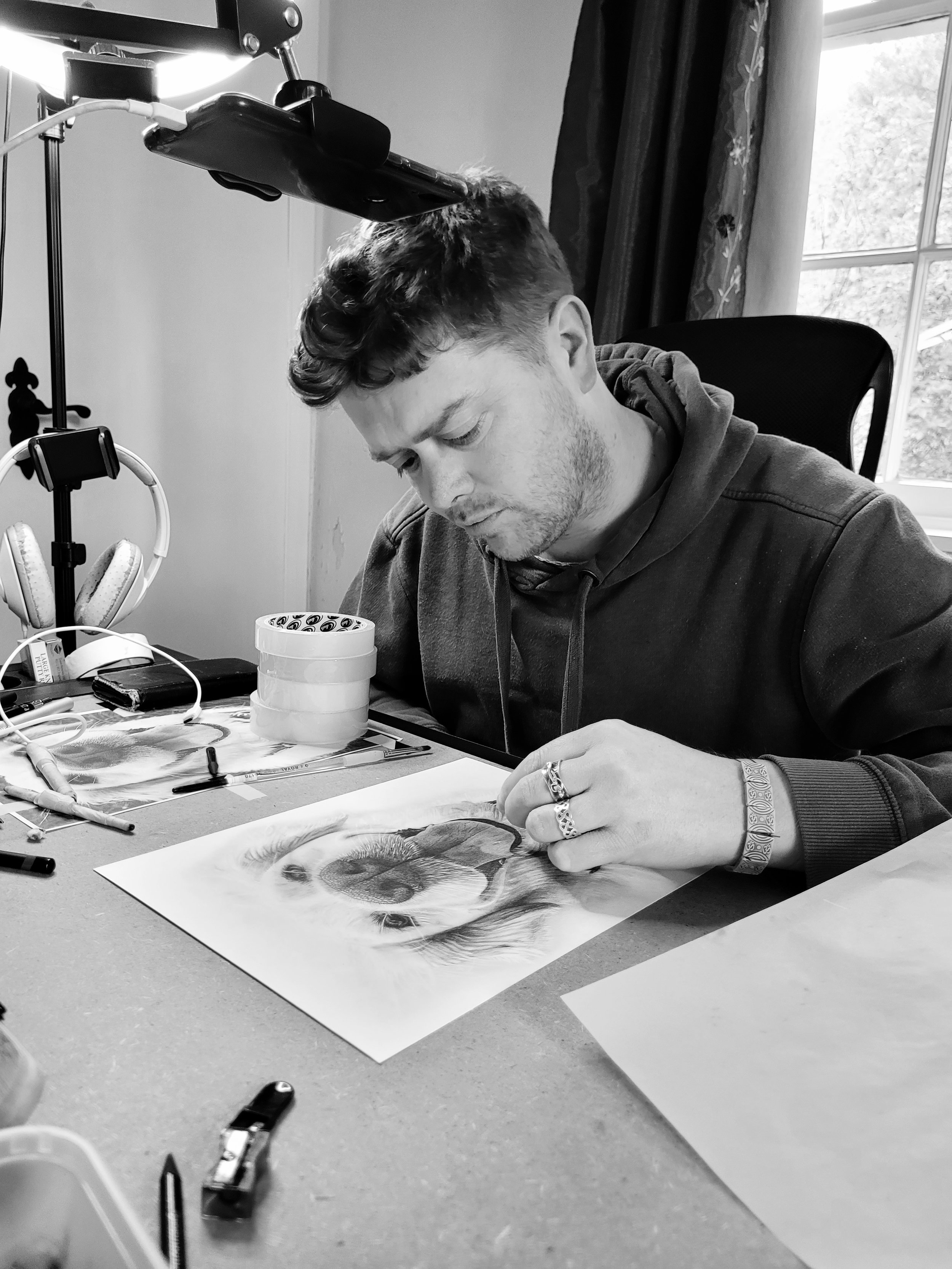 A man working on a detailed pencil drawing of a dog's face at a cluttered desk, with art supplies and headphones nearby, in a room with a window and curtains.