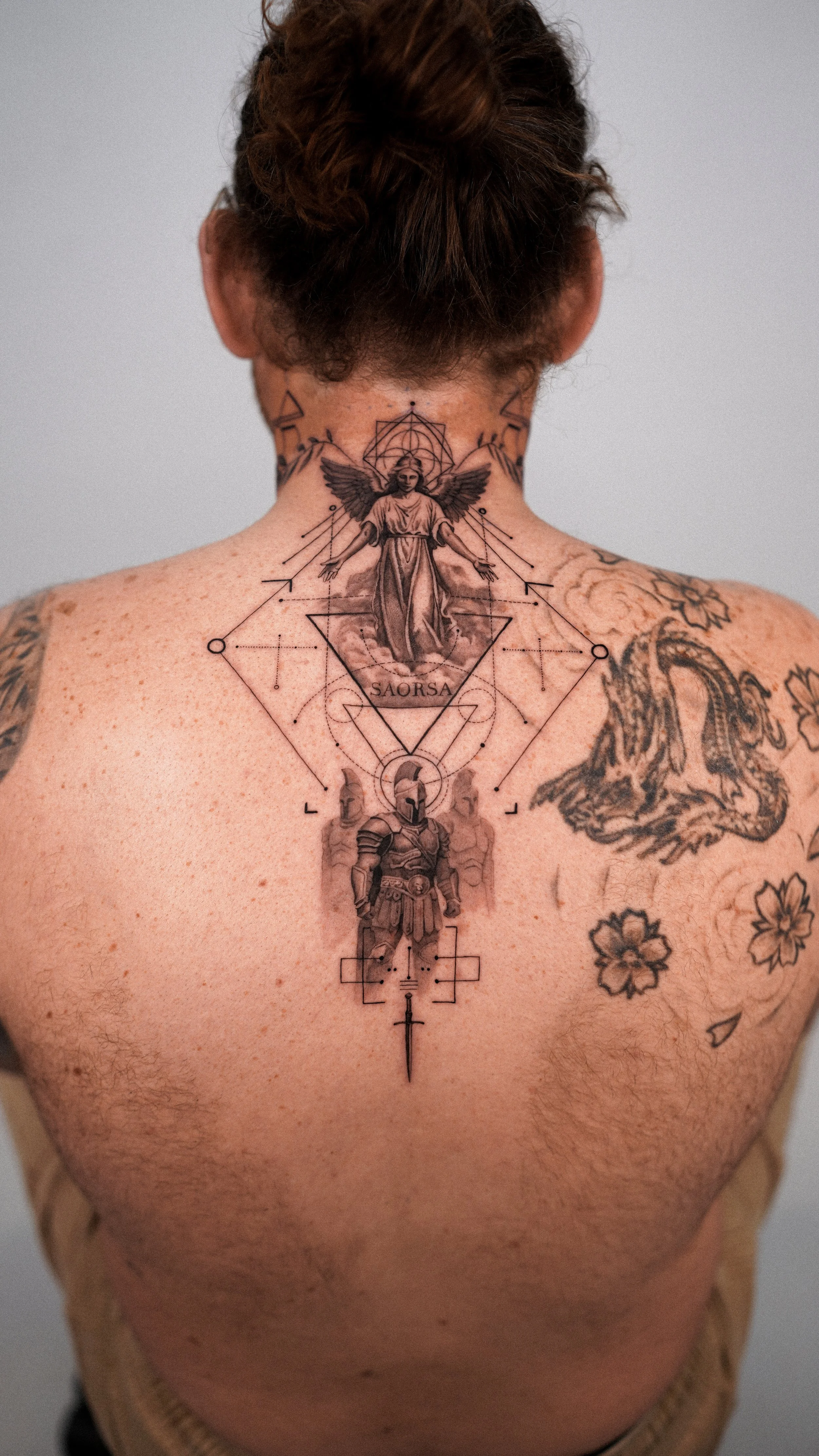 Back view of a person's shoulders and upper back with tattoos, including a large angel tattoo with the word 'SAORSA' and a knight in armor among geometric patterns.