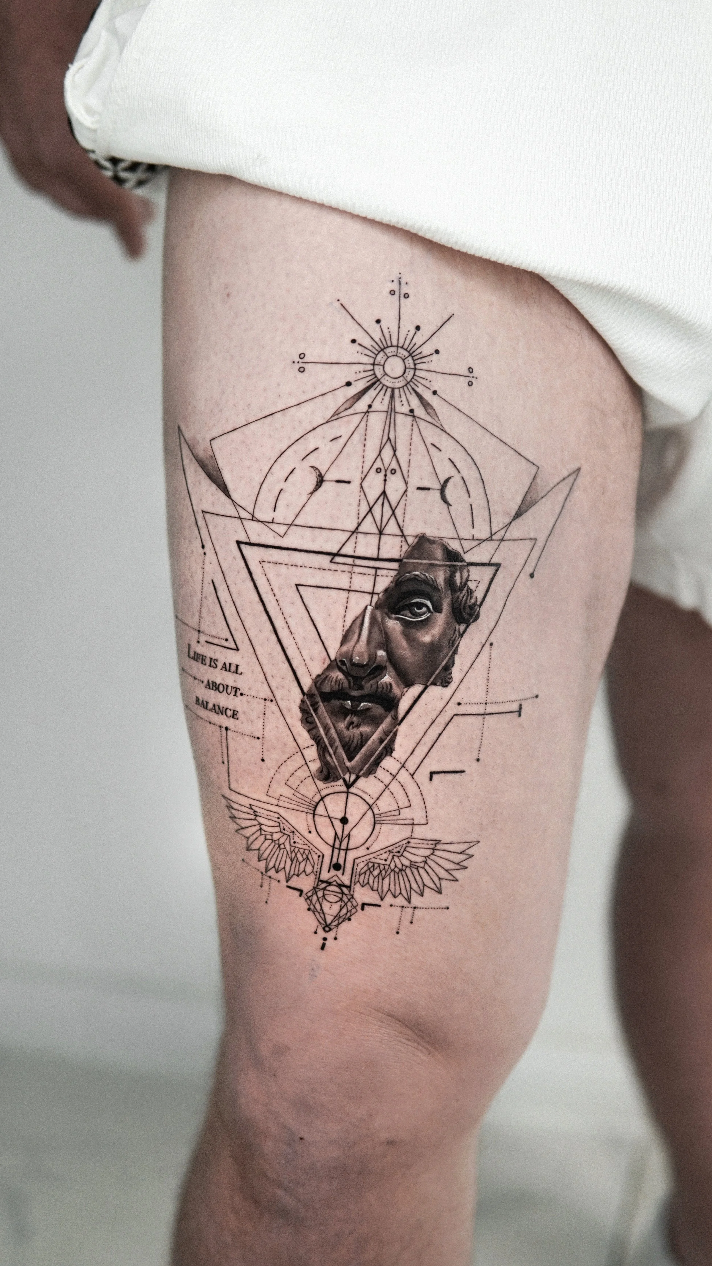 Tattoo on person's thigh featuring geometric lines, a realistic face of an ancient Greek or Roman male with beard and mustache, surrounded by celestial and mystical symbols, with the phrase 'LIFE IS ALL ABOUT BALANCE'.