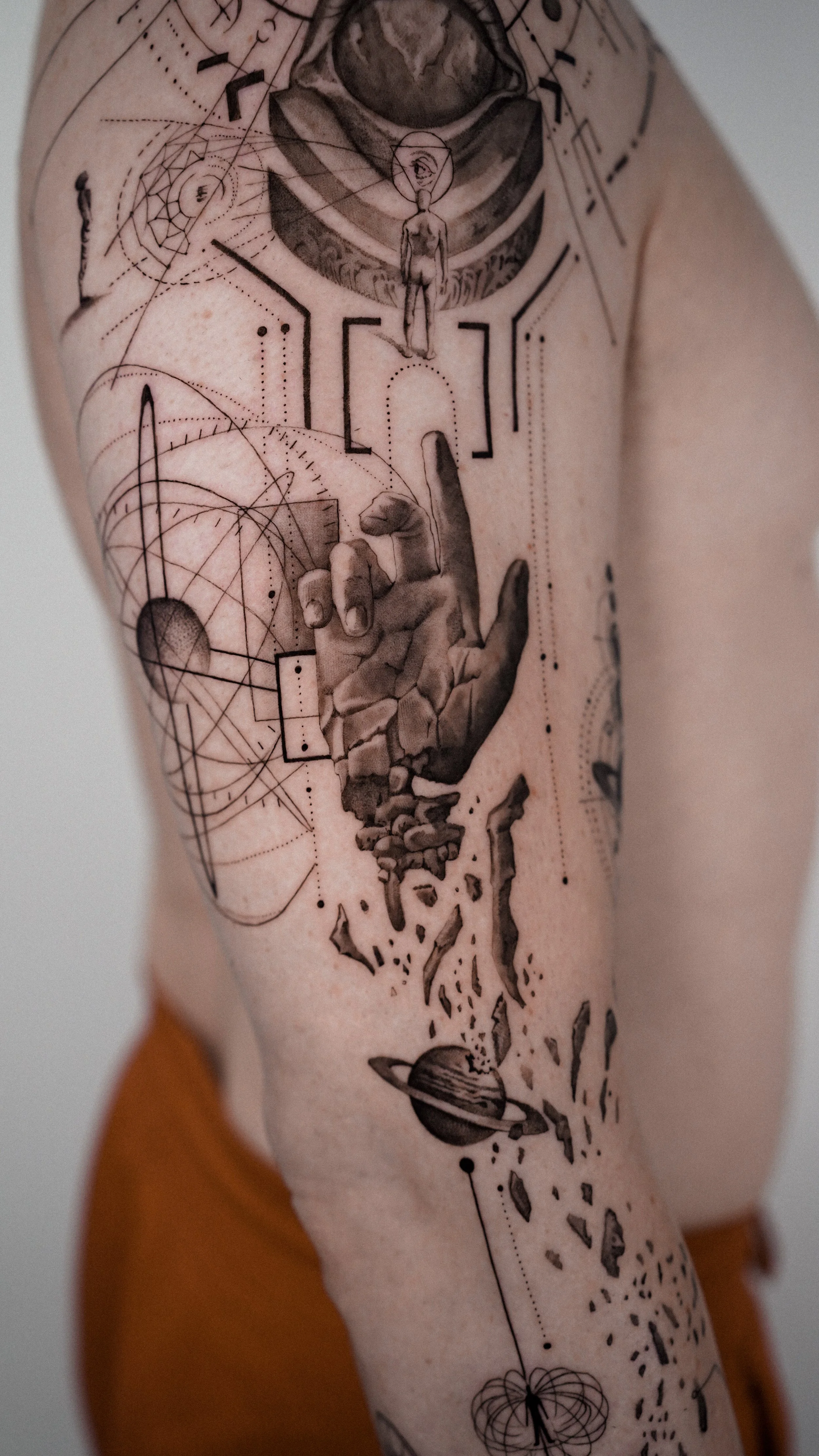 Tattoo on person's arm featuring abstract geometric and cosmic designs, including planets, planets rings, planets, and a human figure with an eye in his head, surrounded by detailed line work and shapes.