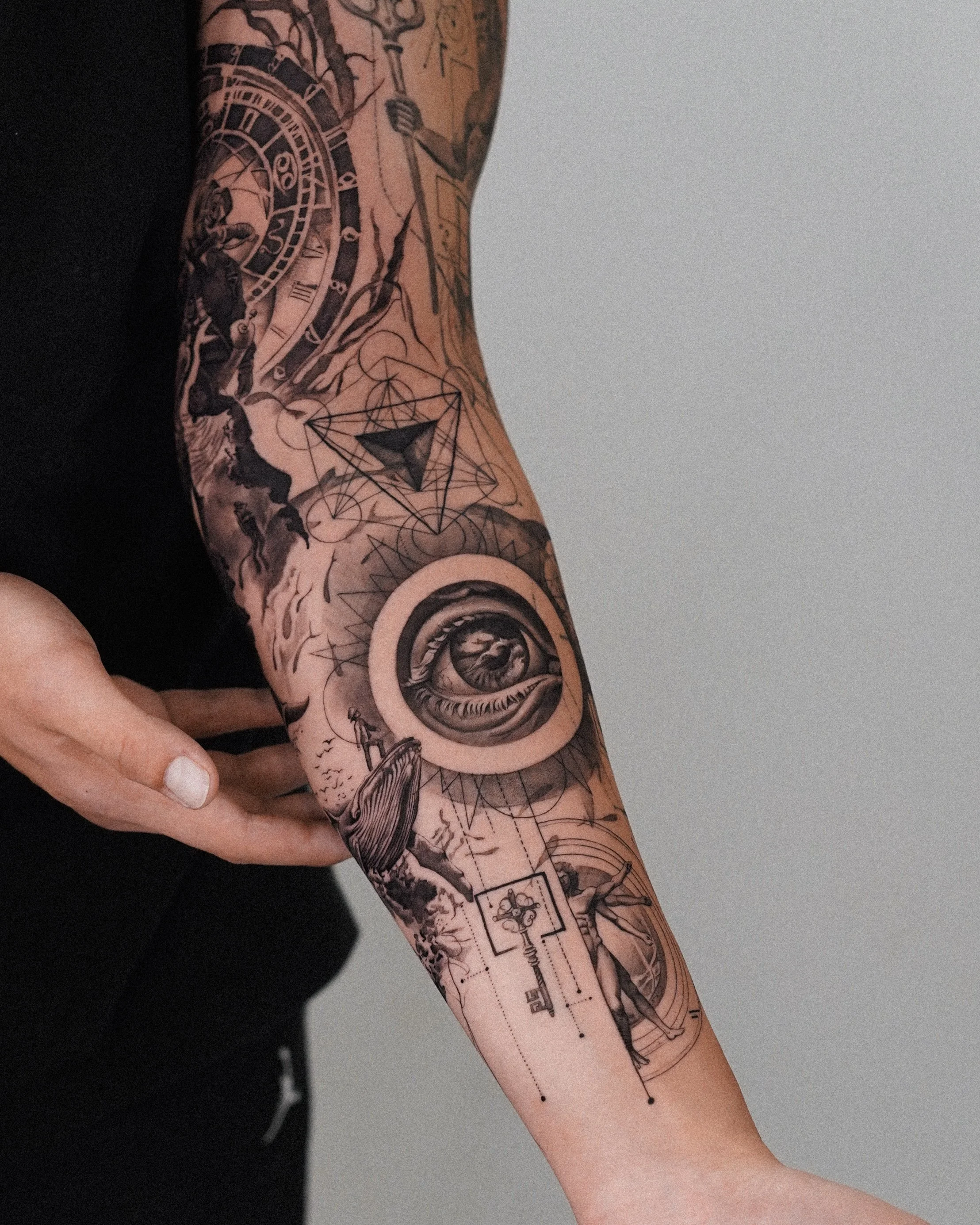 Close-up photo of a forearm with a detailed black and gray tattoo featuring clockwork gears, geometric shapes, and a whale, against a plain background.