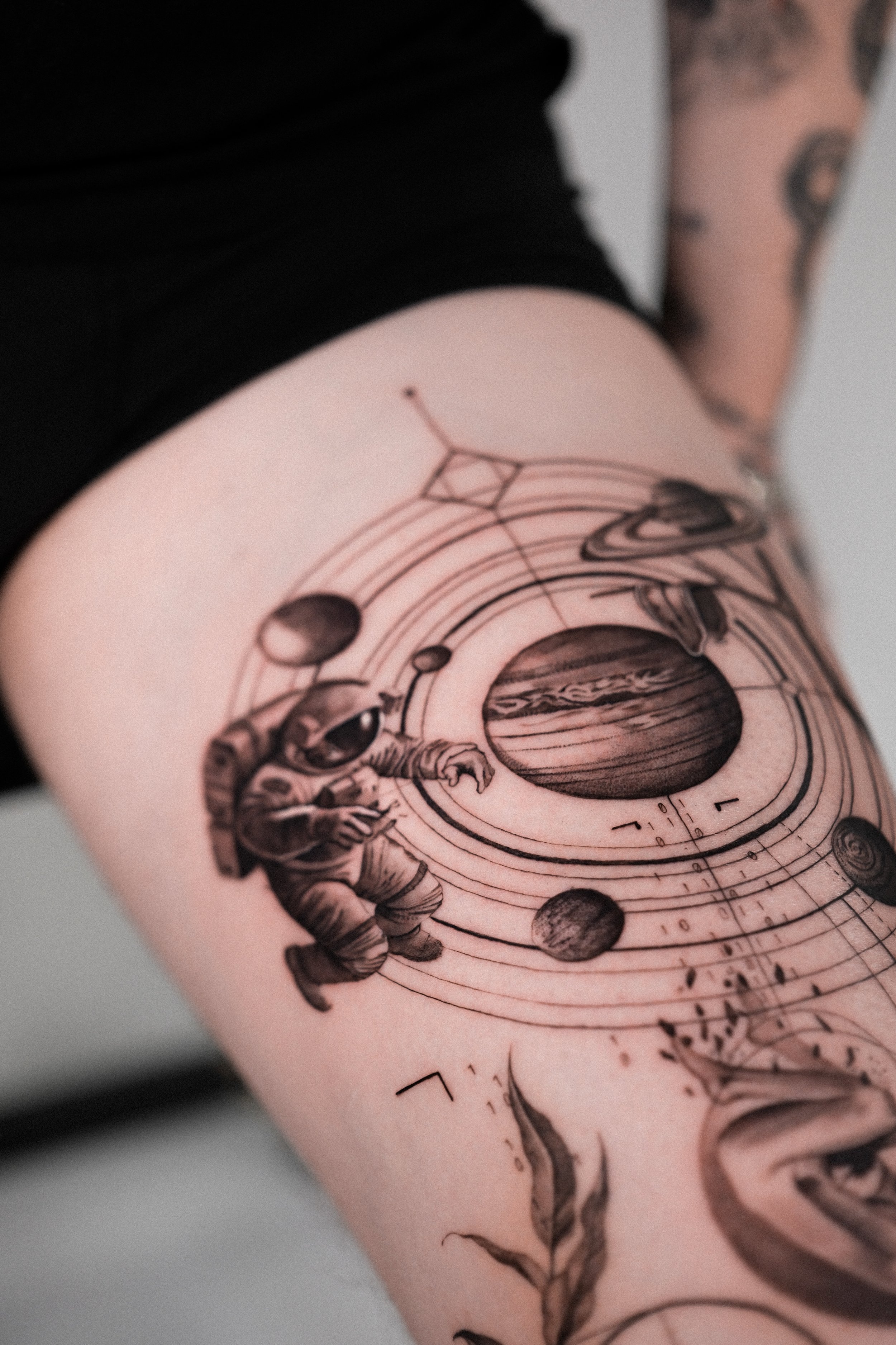 Close-up of a black tattoo on a person's arm, featuring a cosmic scene with planets, a woman, and geometric elements in black ink.