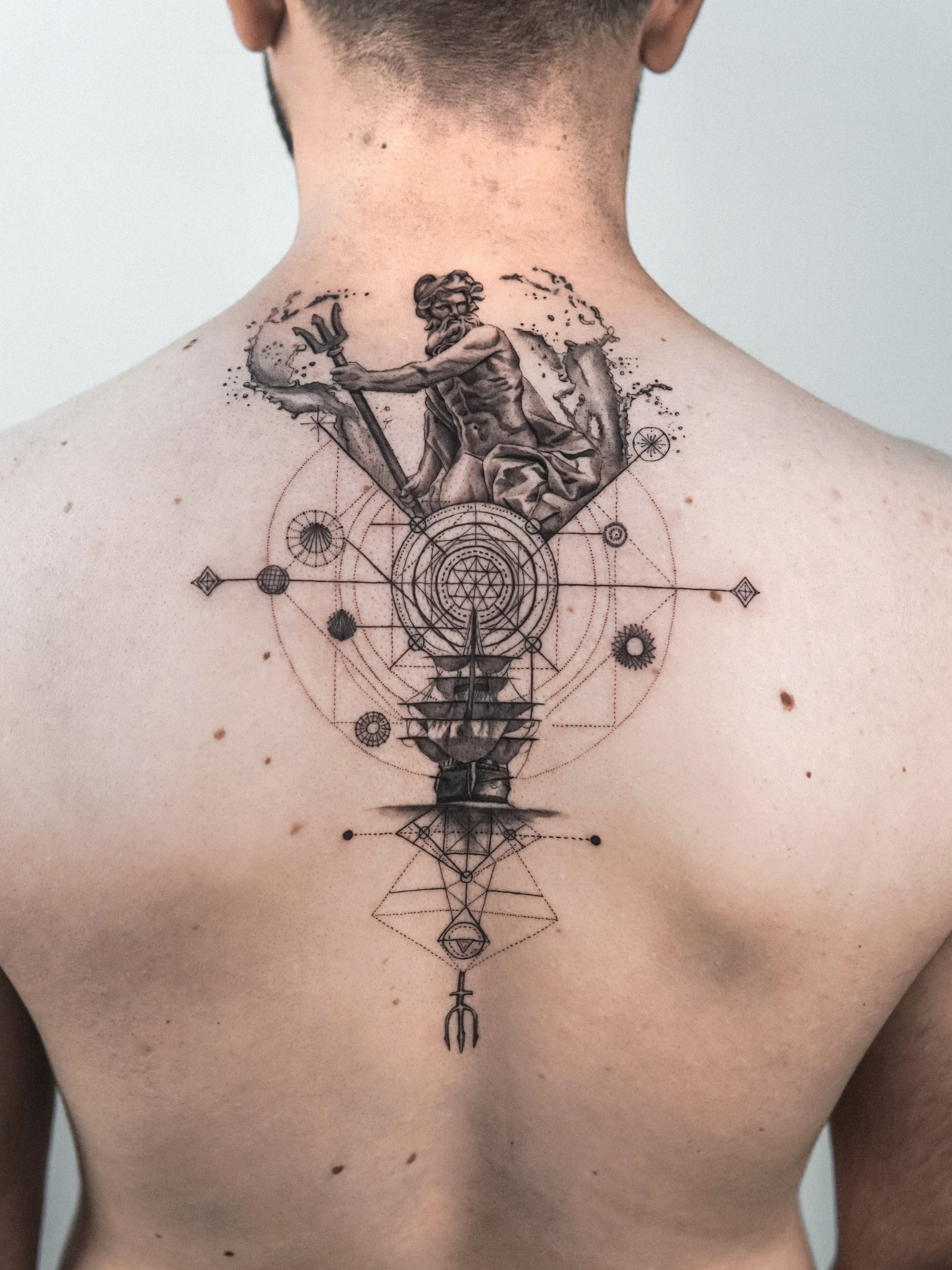 Tattoo on person's chest depicting a muscular ancient Greek god-like figure holding a trident, surrounded by geometric and circular patterns.