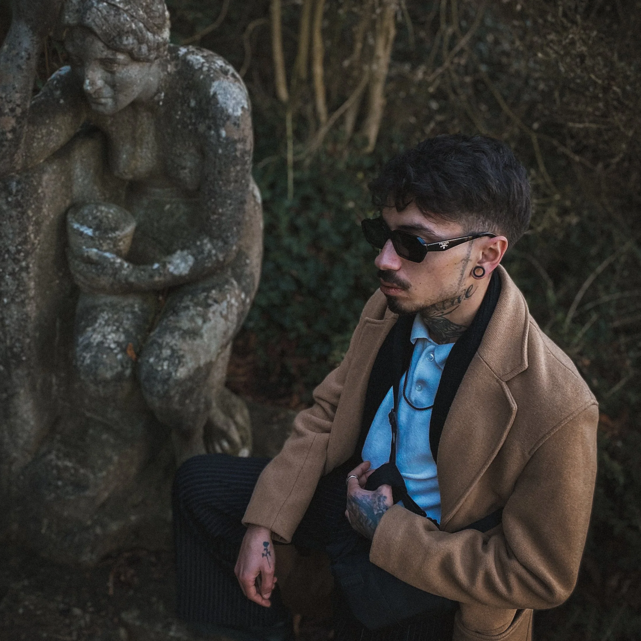 A young man with dark curly hair, sunglasses, and tattoos on his neck, wearing a tan coat and a blue shirt, is standing outside near a stone sculpture of a cherub. The background contains trees and dense foliage.