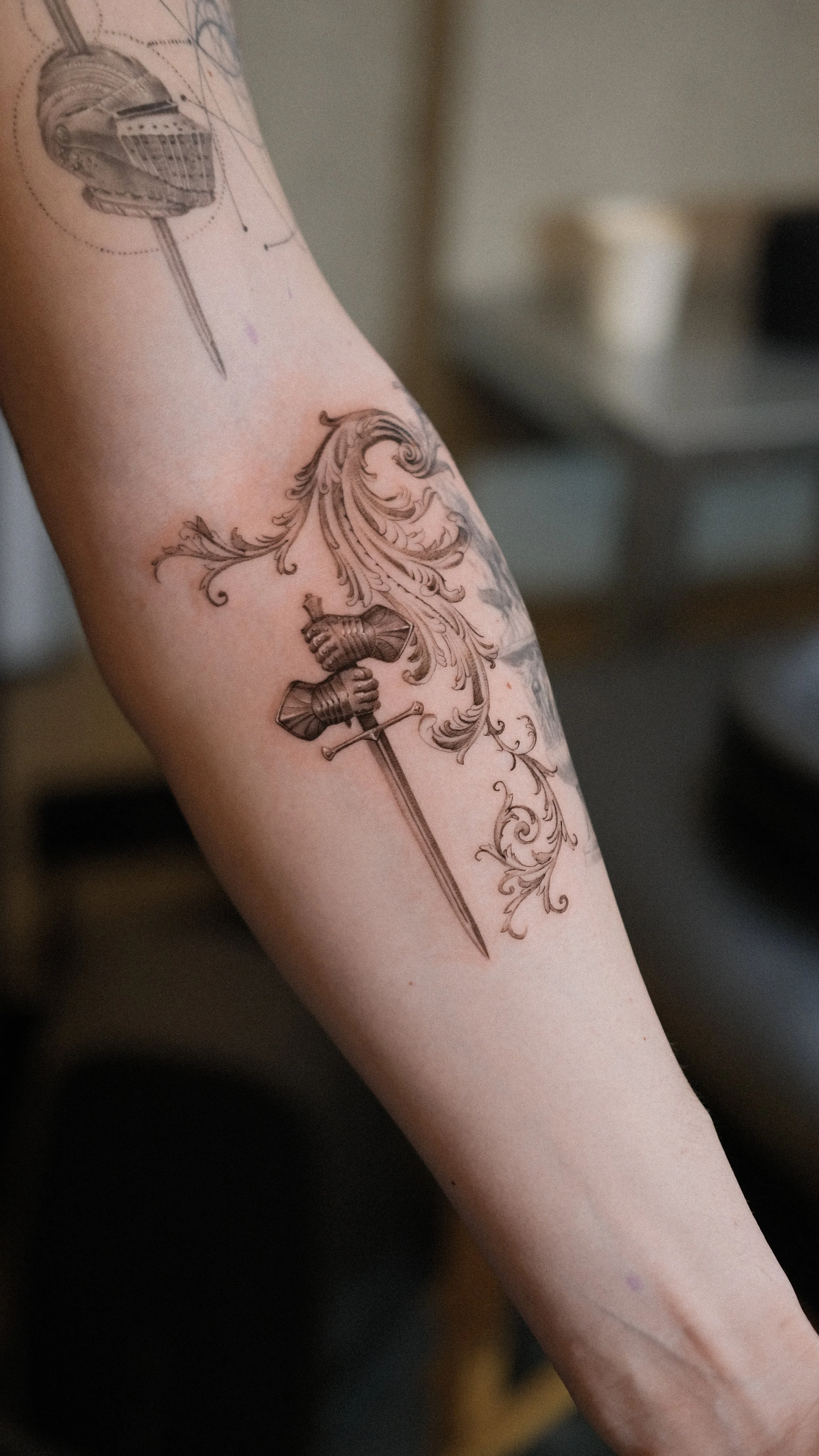 Tattoo of a medieval knight's helmet, ornate swirling floral designs, and a sword on a person's forearm.