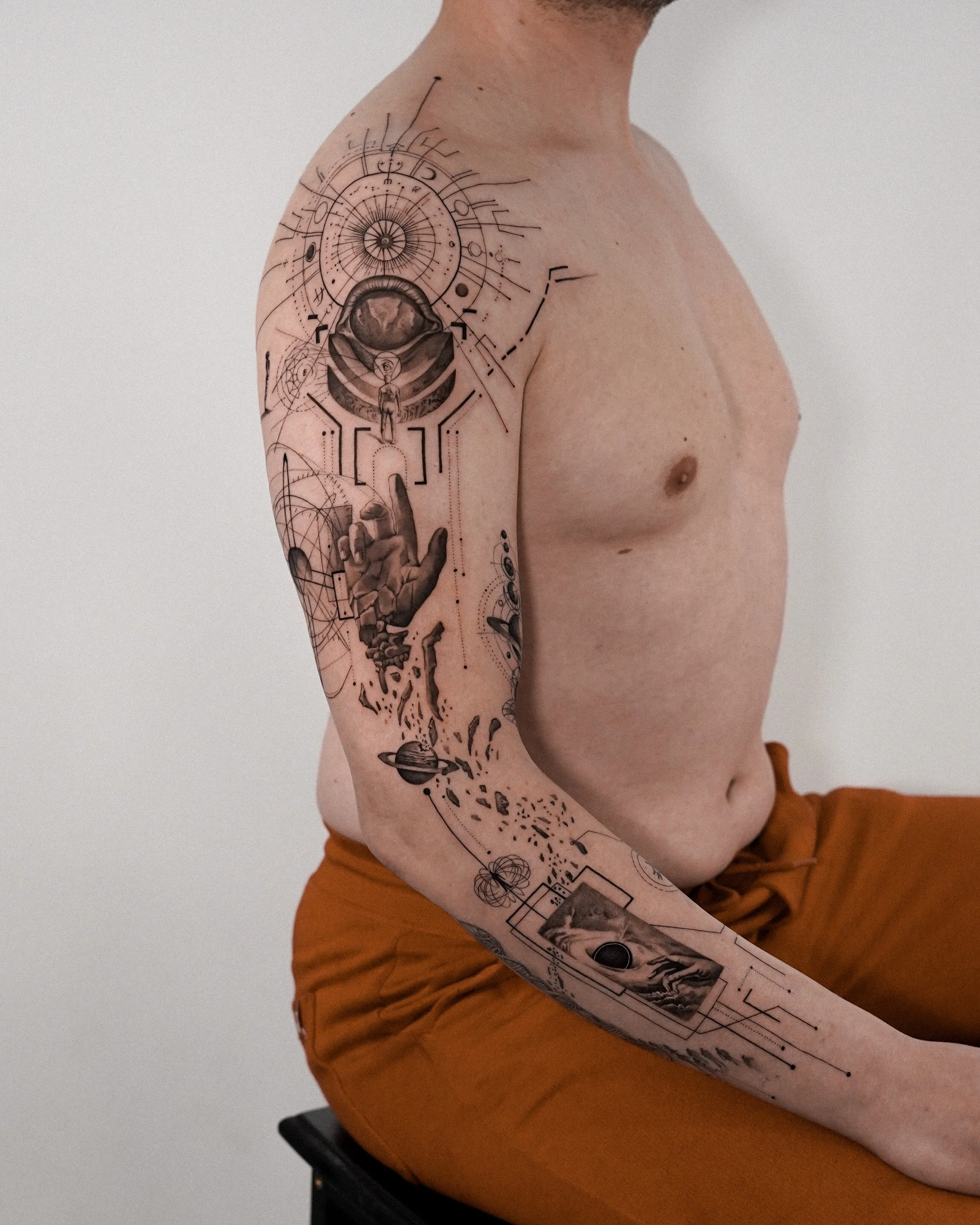 Close-up of a person's left arm and shoulder with a detailed tattoo design. The tattoo features geometric elements, celestial symbols, a human figure, and a landscape scene.