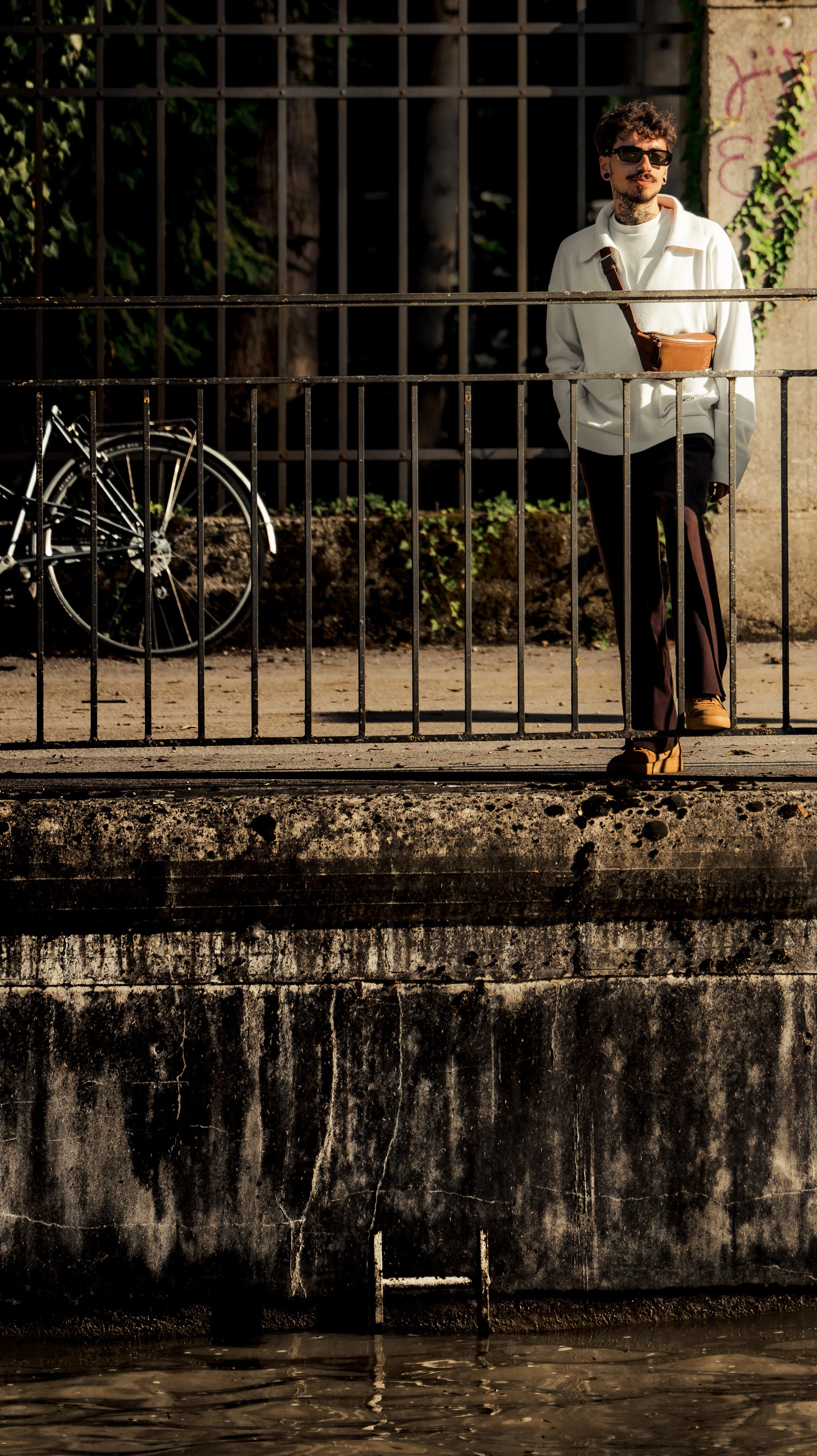 A man with curly hair, sunglasses, and a white jacket stands on a bridge with a metal railing near water. A bicycle is parked against the railing. There is graffiti on the wall behind him and greenery behind the graffiti.