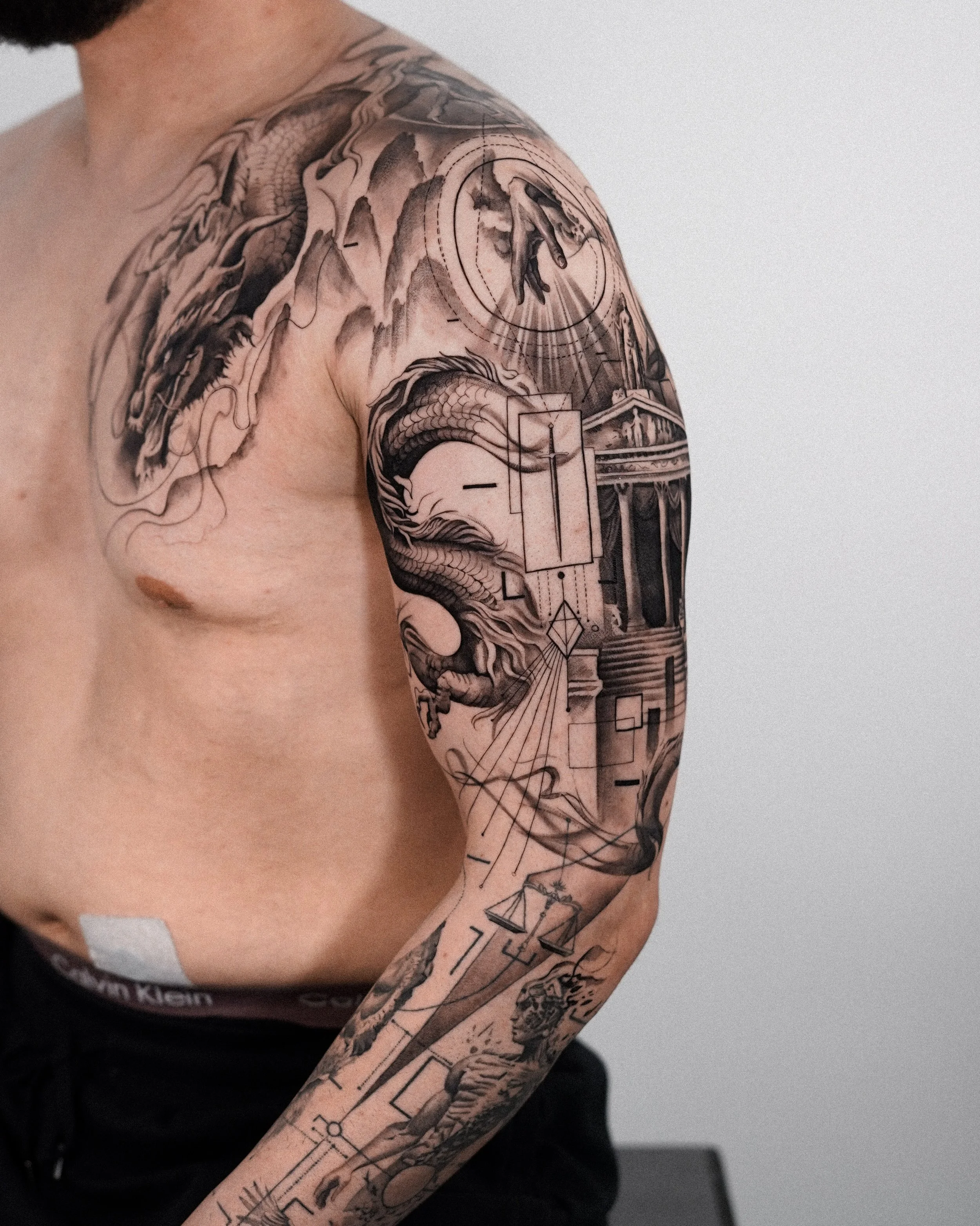 Close-up of a person's upper arm and shoulder, showcasing a detailed black and gray tattoo featuring architectural elements, geometric shapes, shaded clouds, a classical building, and a hand reaching downward.