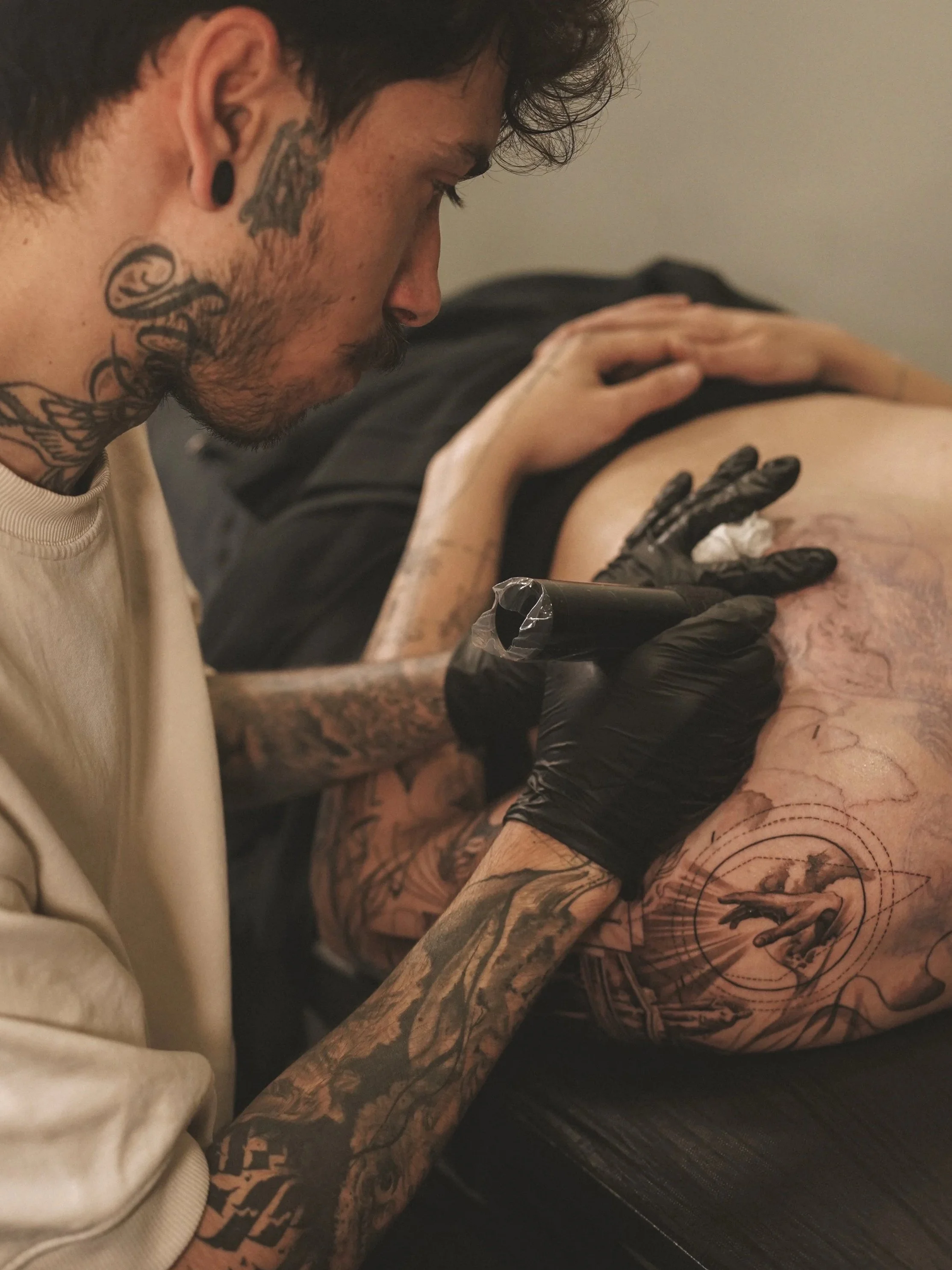 Tattoo artist with dark hair, multiple tattoos on neck and arms, wearing black gloves, working on a tattoo on a person's arm.