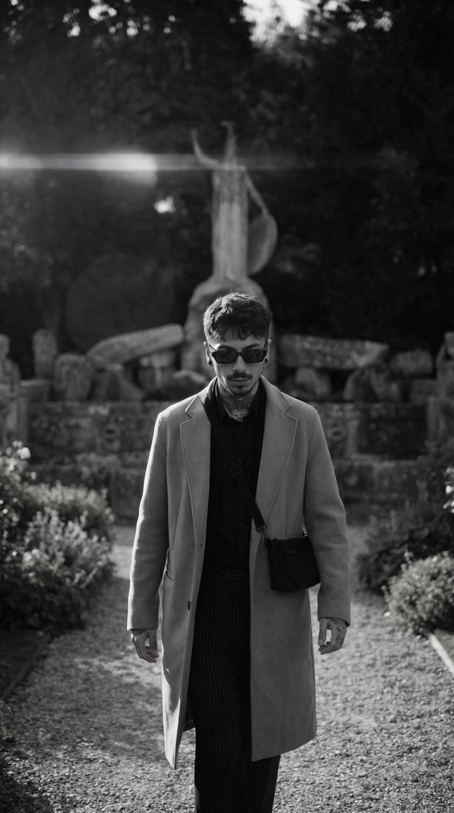 A man with sunglasses, a button-up shirt, and a long coat walking on a garden path with plants on both sides. There is a large snail sculpture and a statue of a woman in the background, with trees behind them.