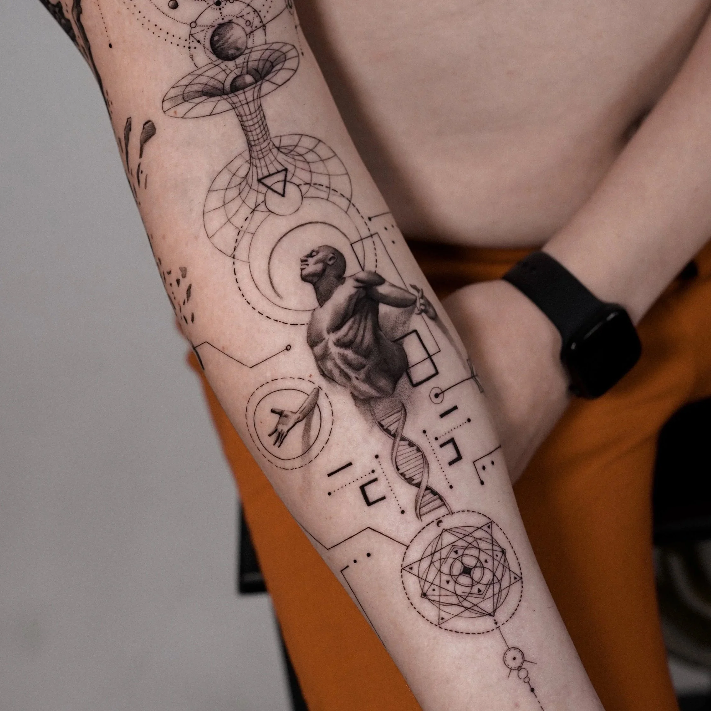Close-up of a person's arm with black geometric sleeve tattoo, including planets, lines, circles, and a lion's head. The skin is light with some visible freckles.