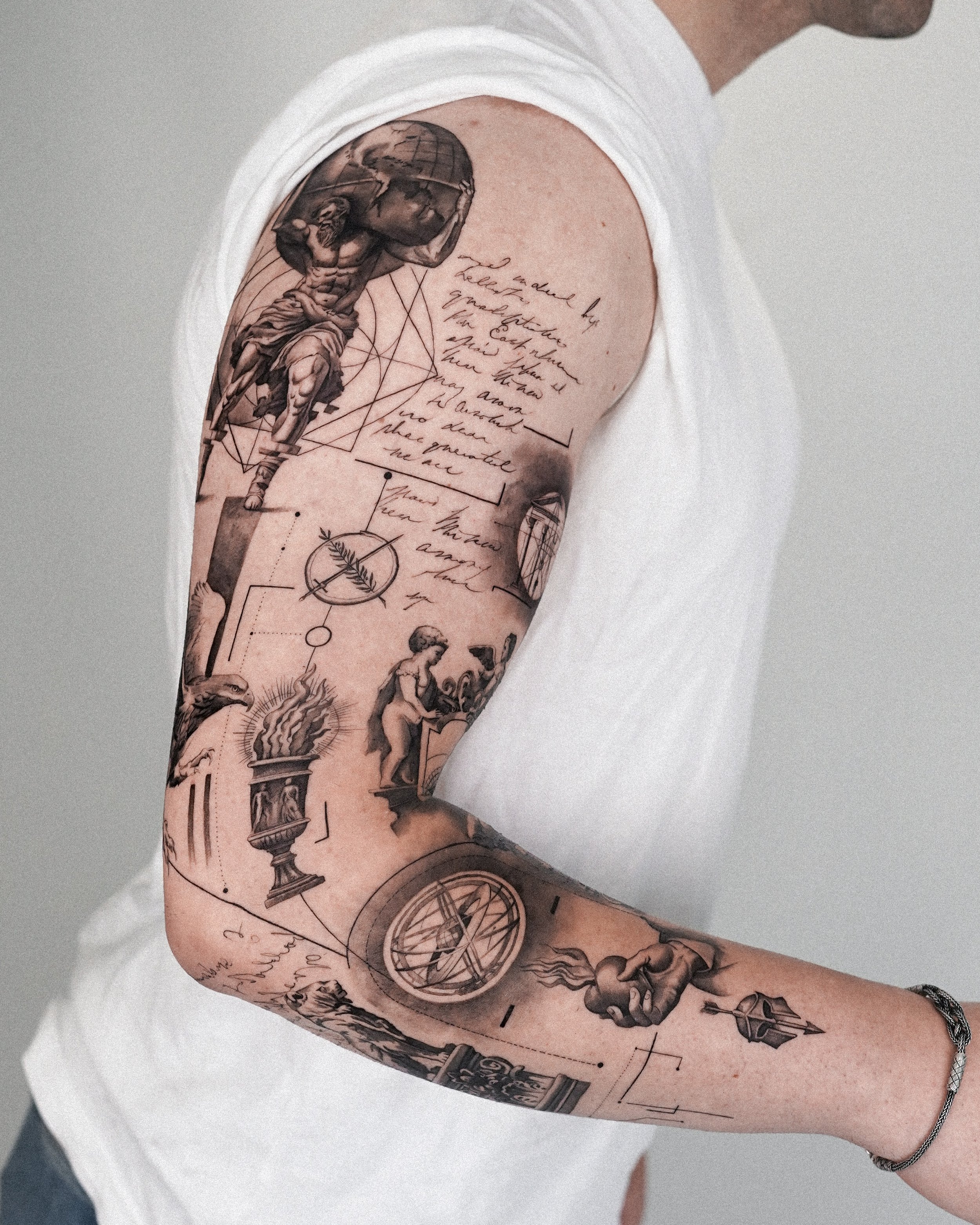 Close-up of a person's arm with a detailed black and gray tattoo sleeve featuring classical art, handwritten text, geometric designs, and decorative elements.