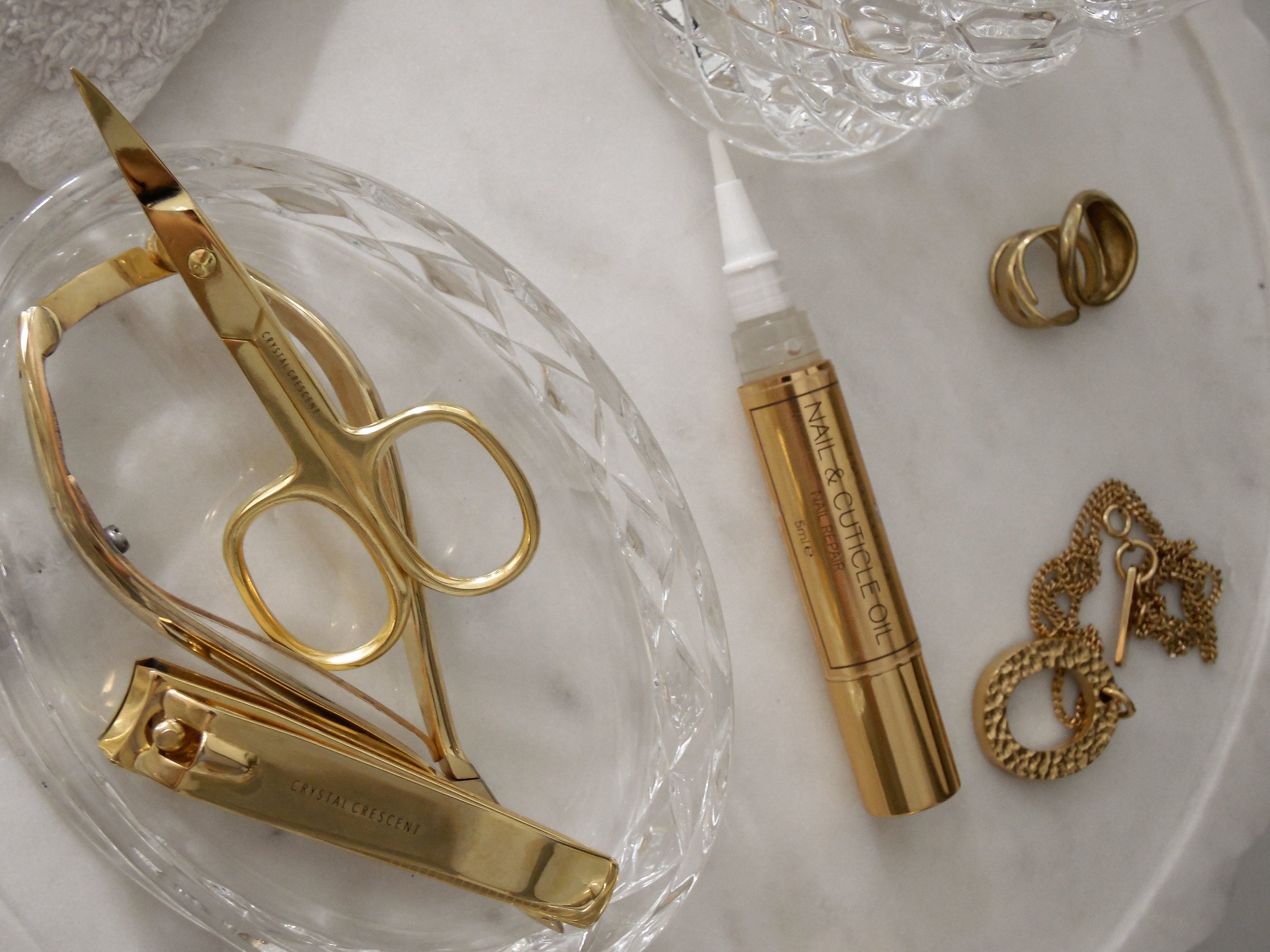 Gold nail and cuticle oil pen, pair of gold hoop earrings, gold scissors, and three gold rings on a white surface with a diamond pattern glass container.