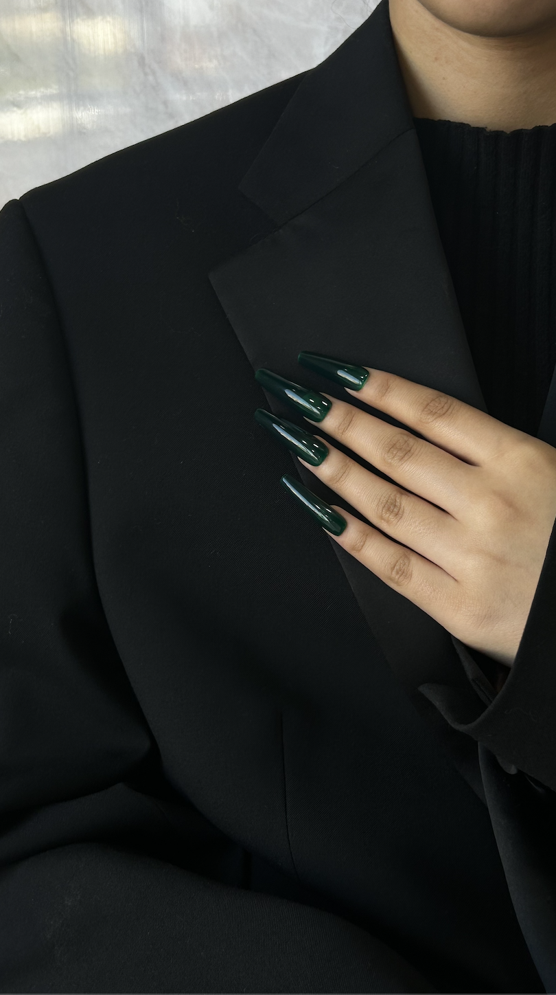 Close-up of a person's hand with long dark green manicured nails resting on their black blazer, with part of their neck and black top visible.