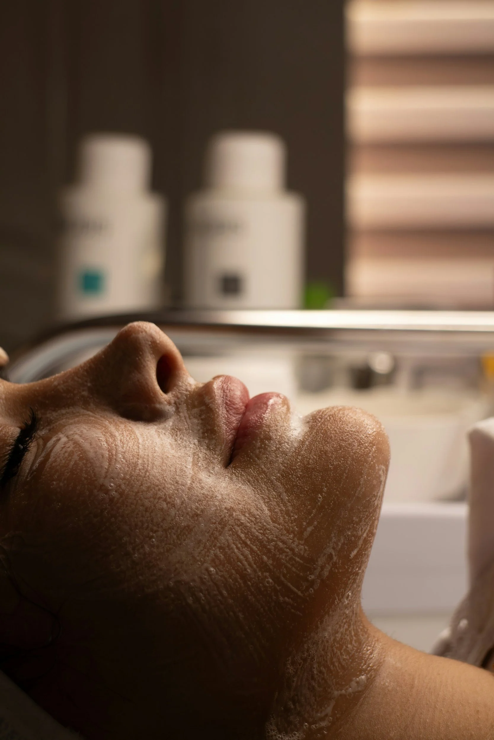Close-up of a woman receiving facial treatment with soap or cleanser on her face, lying down with bottles in the background.