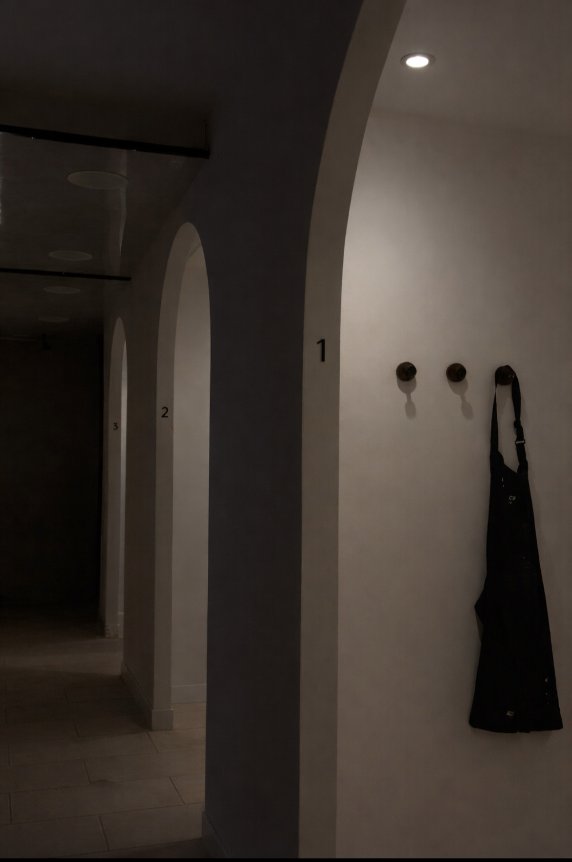 Interior hallway with numbered archways, hooks with a black apron hanging on the wall, and a ceiling spotlight.