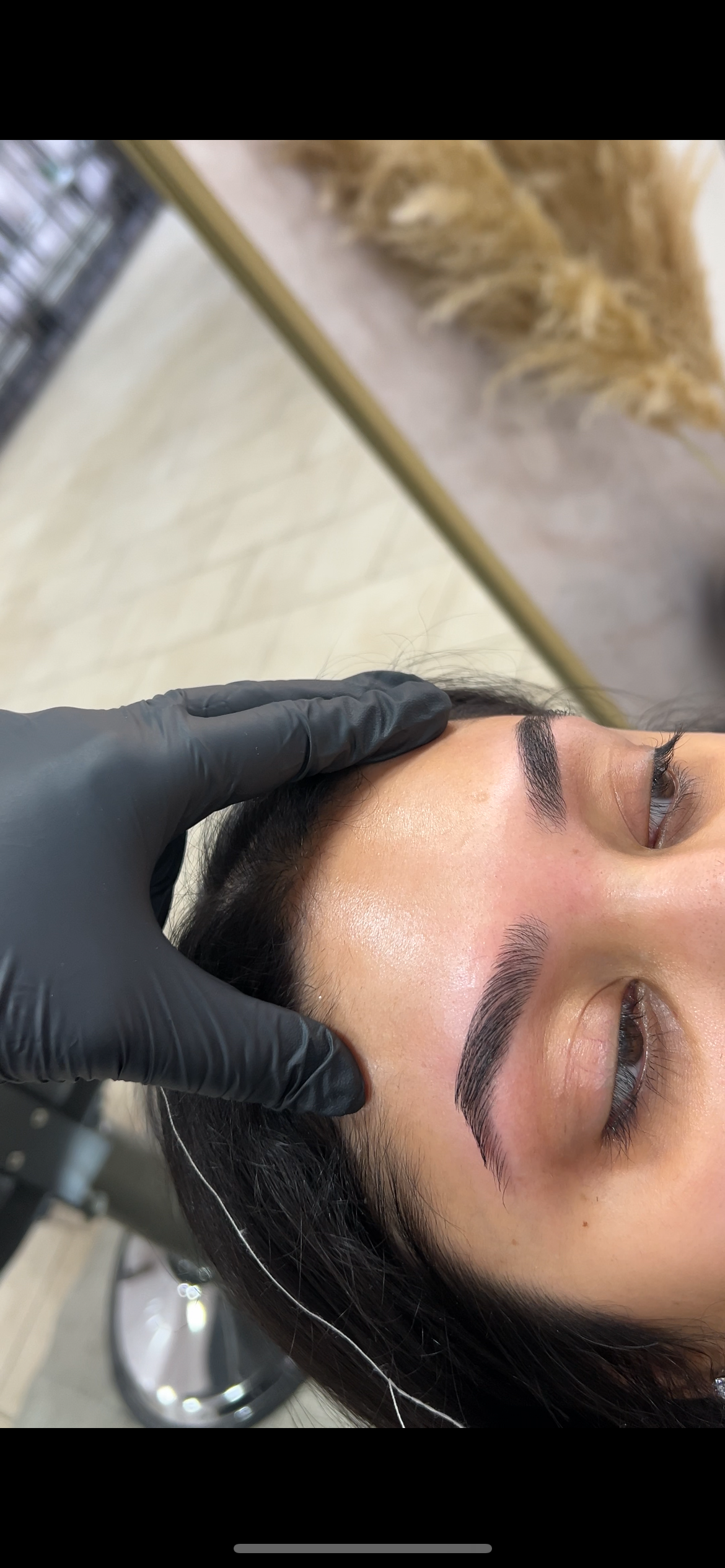Close-up of a woman receiving an eyebrow treatment at a salon, with a gloved hand gently holding her forehead. The woman has dark hair and well-groomed eyebrows.