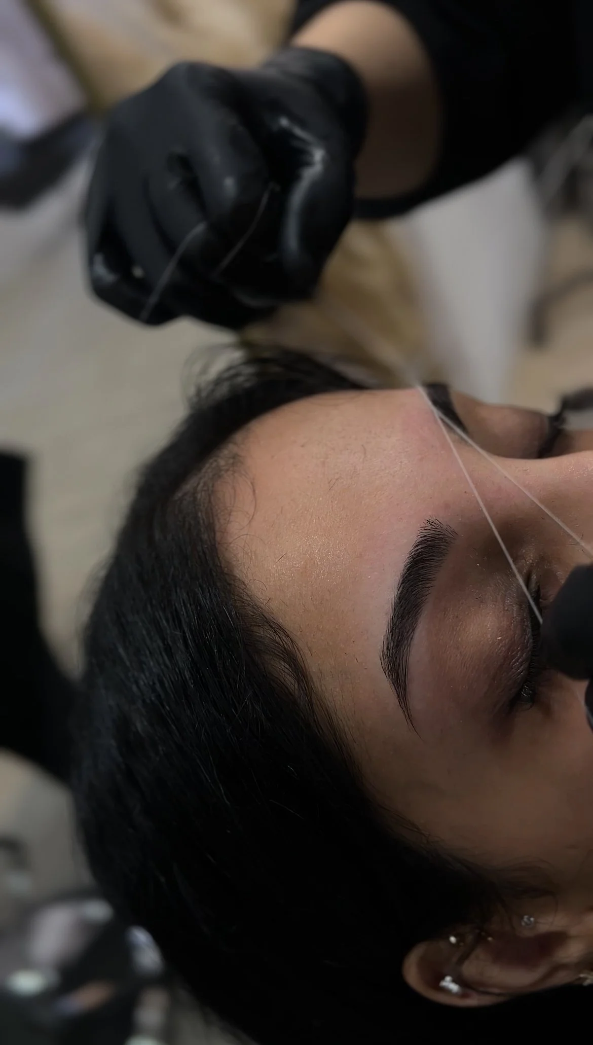A woman receiving a cosmetic procedure on her eyebrows using a thread, with her eyes closed.