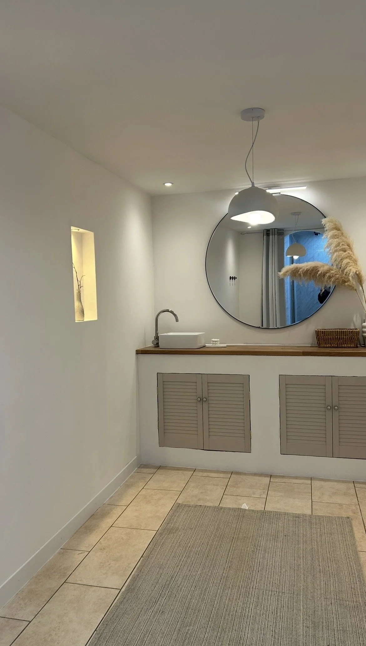 Minimalist bathroom with a large round mirror, a white vessel sink, and a wall-mounted faucet. A pendant light hangs above the sink. A small rectangular niche with a vase inside is built into the wall. The vanity cabinet has two louvered doors. A pam