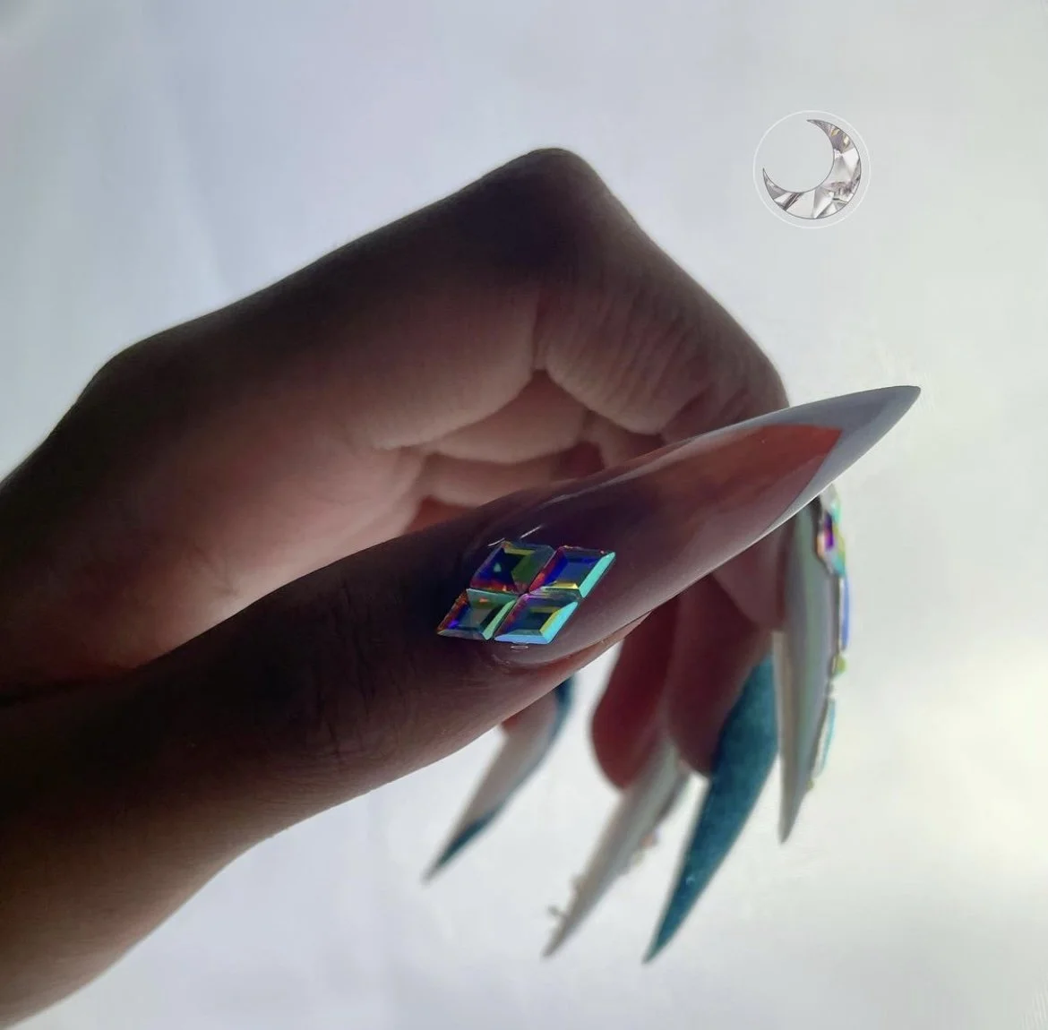 Close-up of a hand with decorated fingernails, holding a card with holographic stickers, and a white background with a small decorative moon in the top right corner.