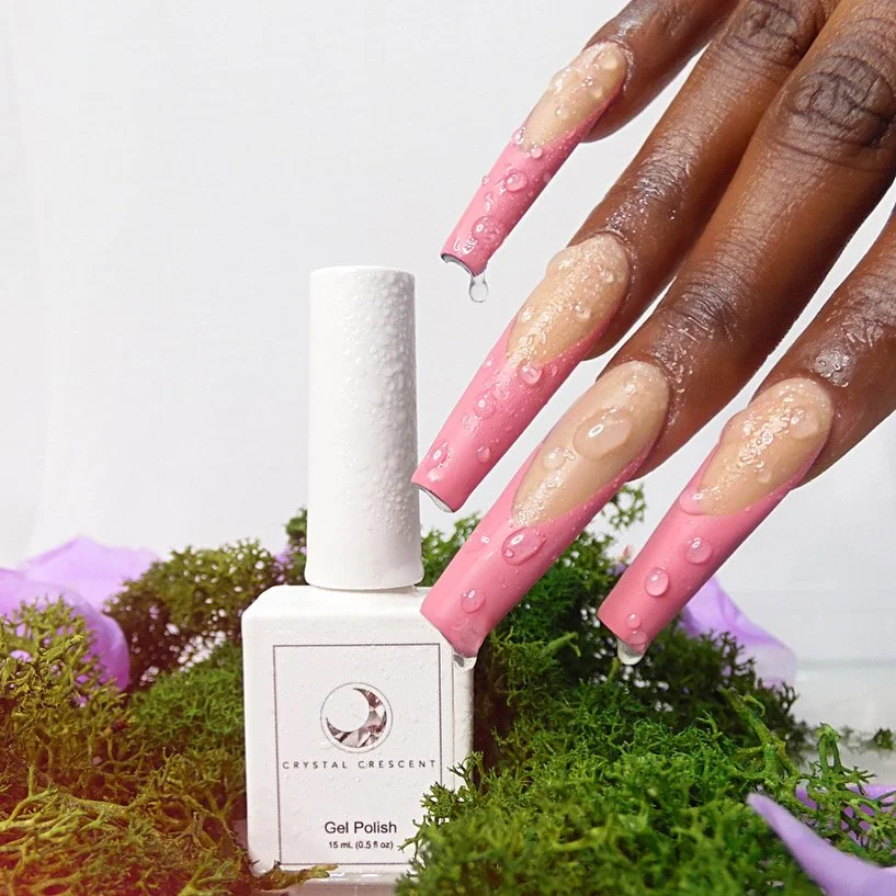 A person's hand with freshly painted long, coffin-shaped nails in pink with beige tips, touching the cap of a white bottle of gel nail polish surrounded by green moss and purple flowers.