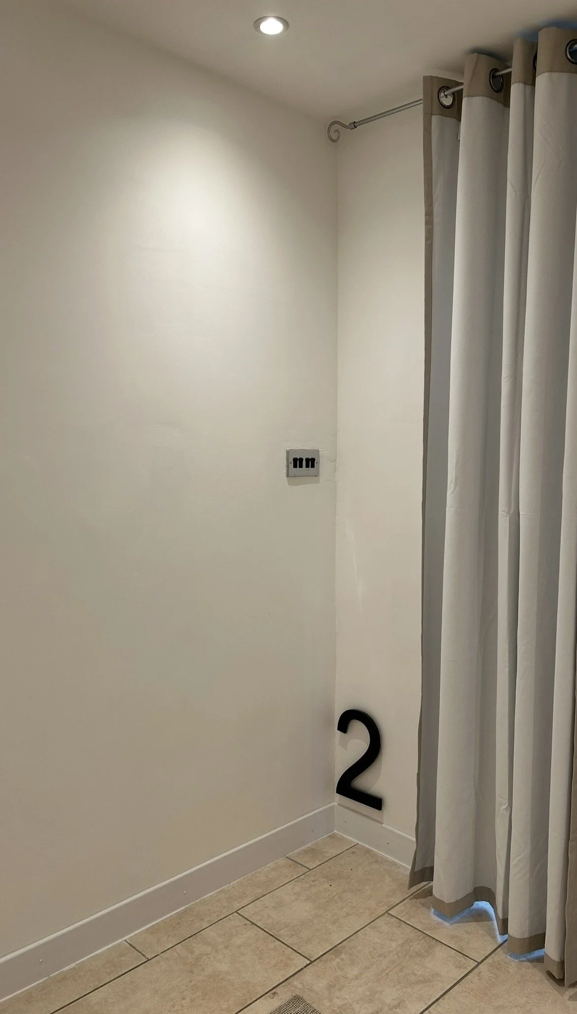 Corner of a room with a white wall, beige tiled floor, a wall outlet, a number 2 on the wall near the baseboard, and a gray and white curtain hanging from a metal rod.