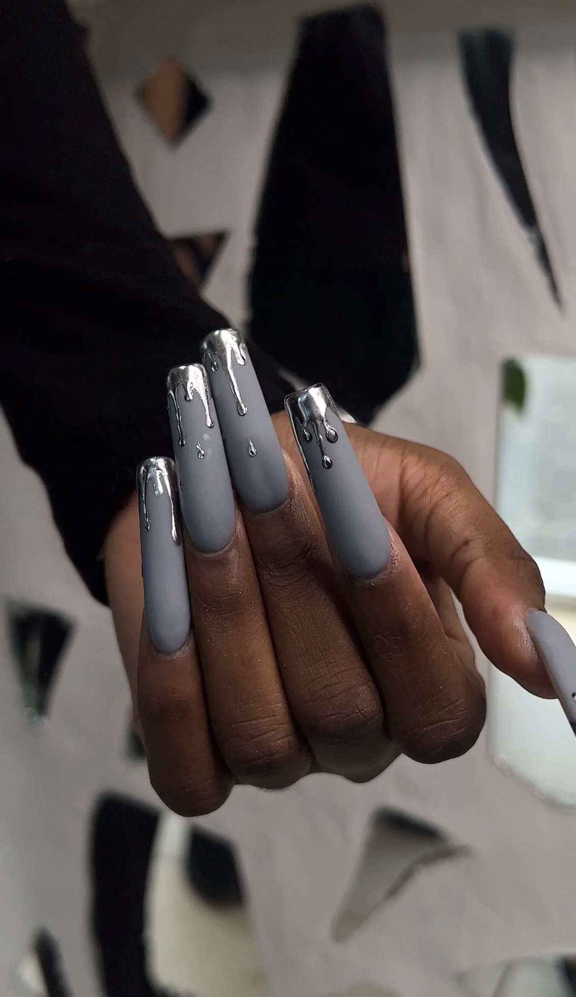 Close-up of a hand with long, coffin-shaped nails painted with a matte gray polish and silver metallic drip design.