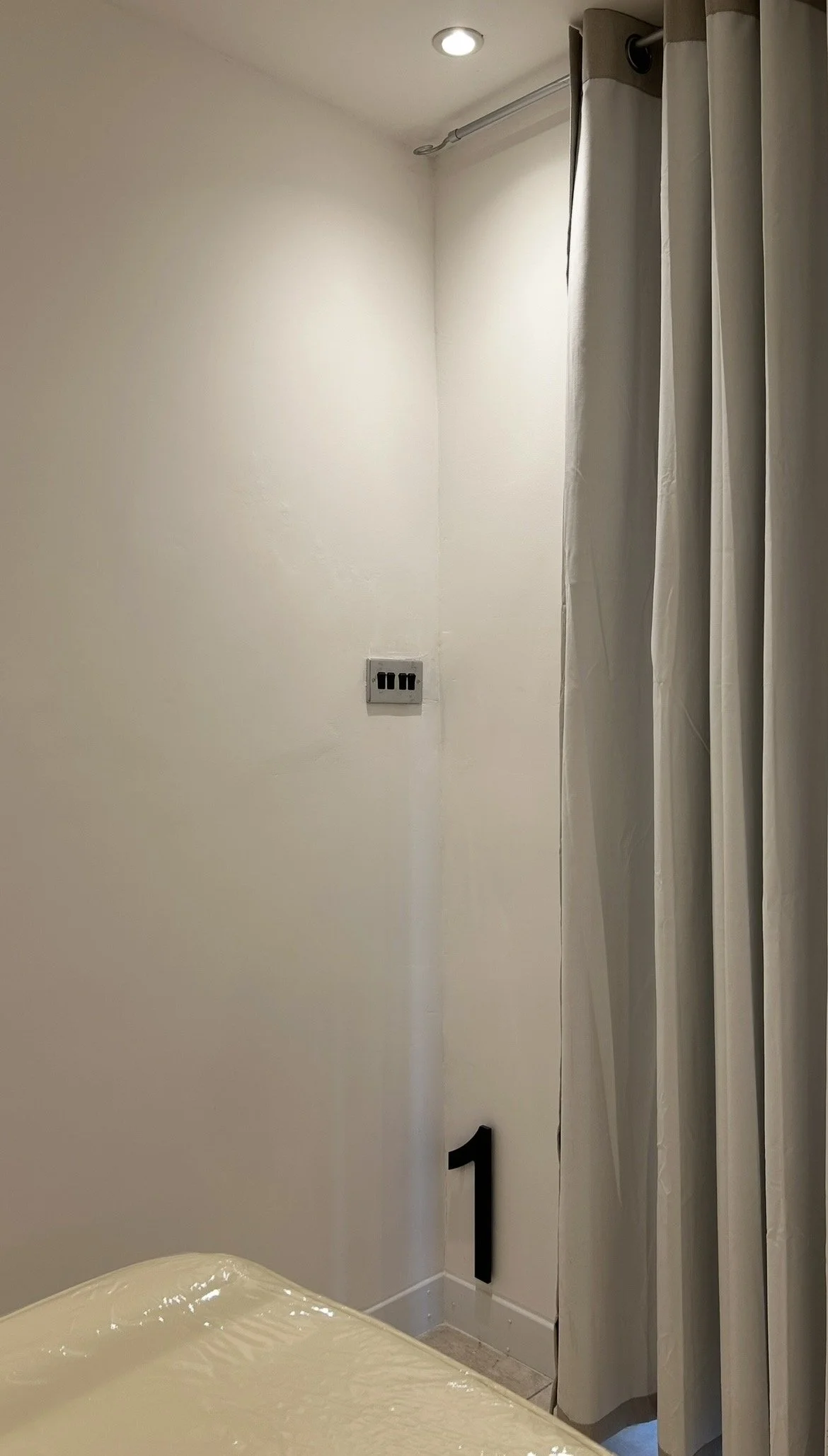 Empty room with white walls, a window with closed blinds, and a dark decorative '1' on the floor near the corner.
