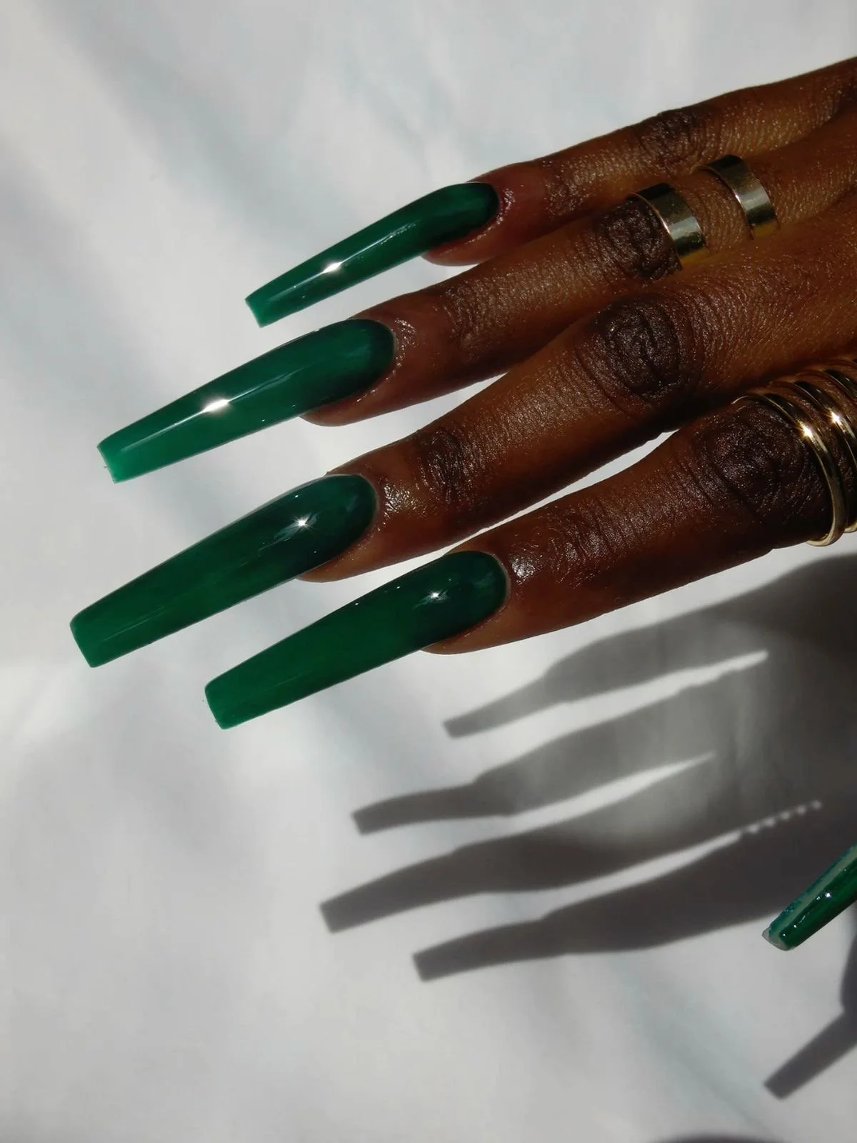 Hand with long green acrylic nails, multiple gold rings and rings on fingers, casting shadows on a white surface.