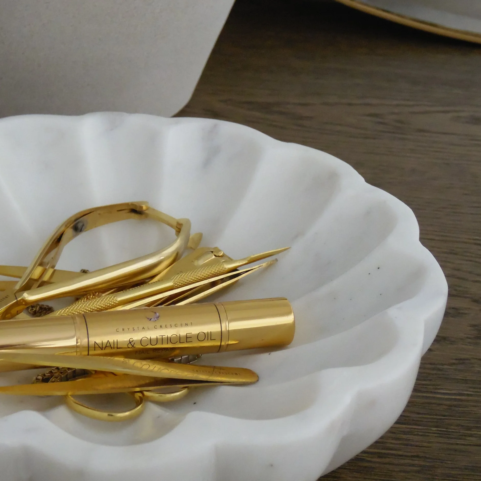A white ceramic dish holding gold nail and cuticle tools, including a nail clipper, cuticle pushers, and a nail oil pen, placed on a wooden surface.