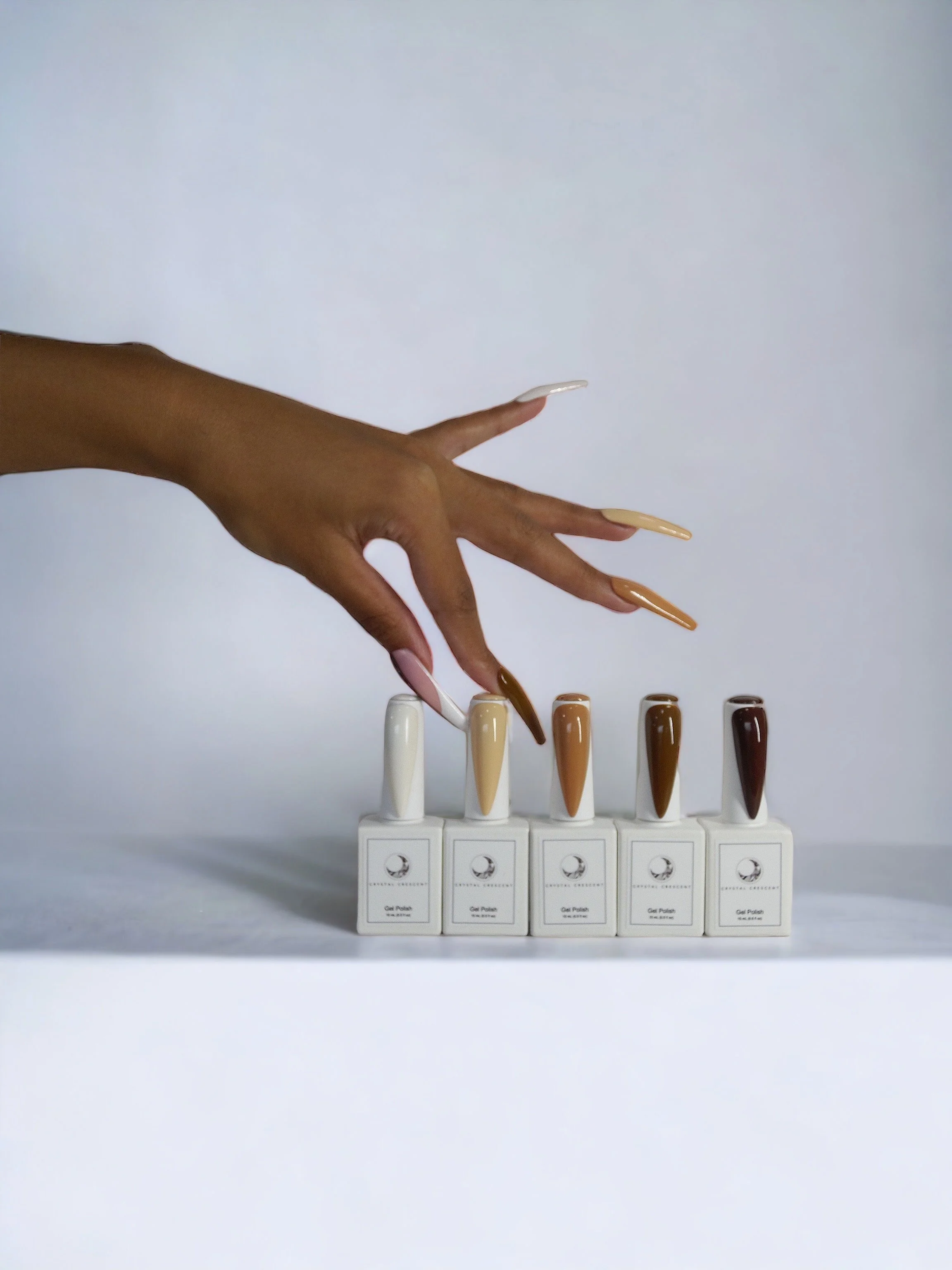 Hand with manicured nails reaching over bottles of gel nail polish in different shades of beige and brown, arranged on a white surface with a white background.
