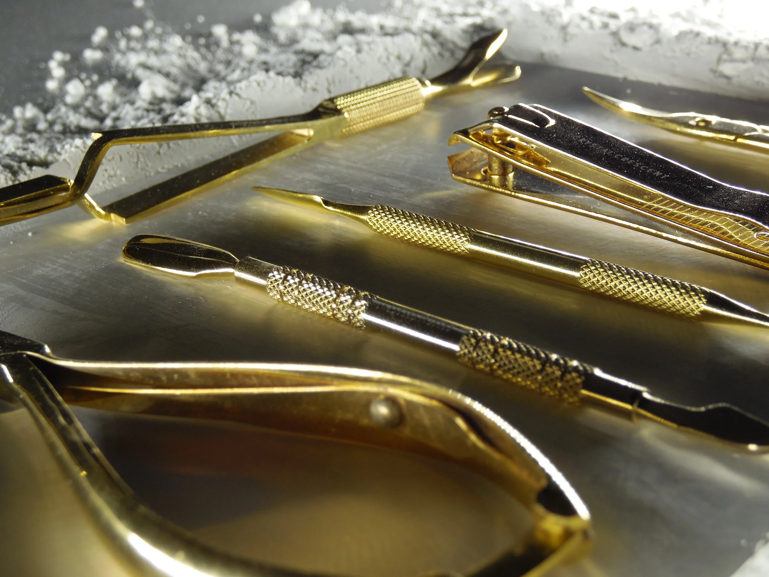 Set of gold-colored manicure tools including nail scissors, cuticle nippers, and nail files arranged on metallic surface.