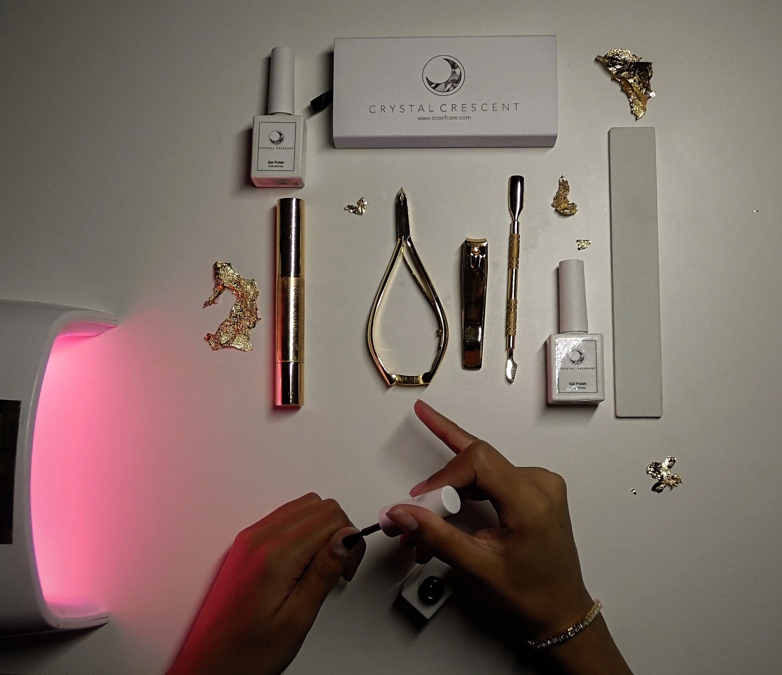 A flat lay of various beauty tools and products on a white surface, including nail polish, a cuticle nipper, a nail file, a tool with a pointed end, small gold decorative pieces, and a box labeled 'Crystal Crescent'.