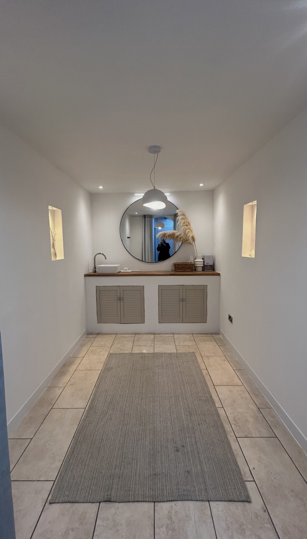 A minimalistic hallway with a tiled beige floor, a grey rug, and white walls. At the end, there is a small vanity area with a round mirror, a wooden countertop, a white basin sink, and some decor and toiletries. The hallway has small decorative niche