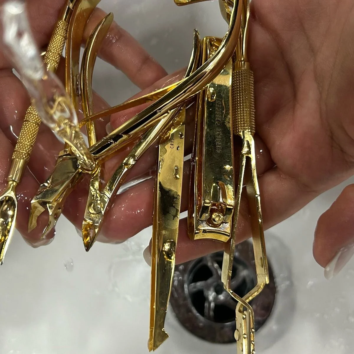 Close-up of a hand holding multiple gold-colored crystal crescent nail tools under running water in a sink.