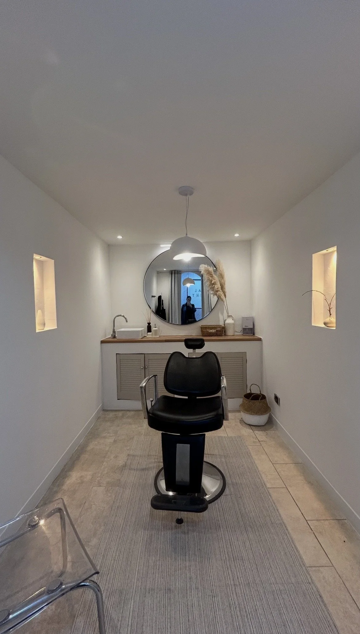 A modern salon interior with a black salon chair facing a wooden counter with a round mirror, interior lighting, decorative vases, and pampas grass arrangements. There are small wall niches with lighting on either side.