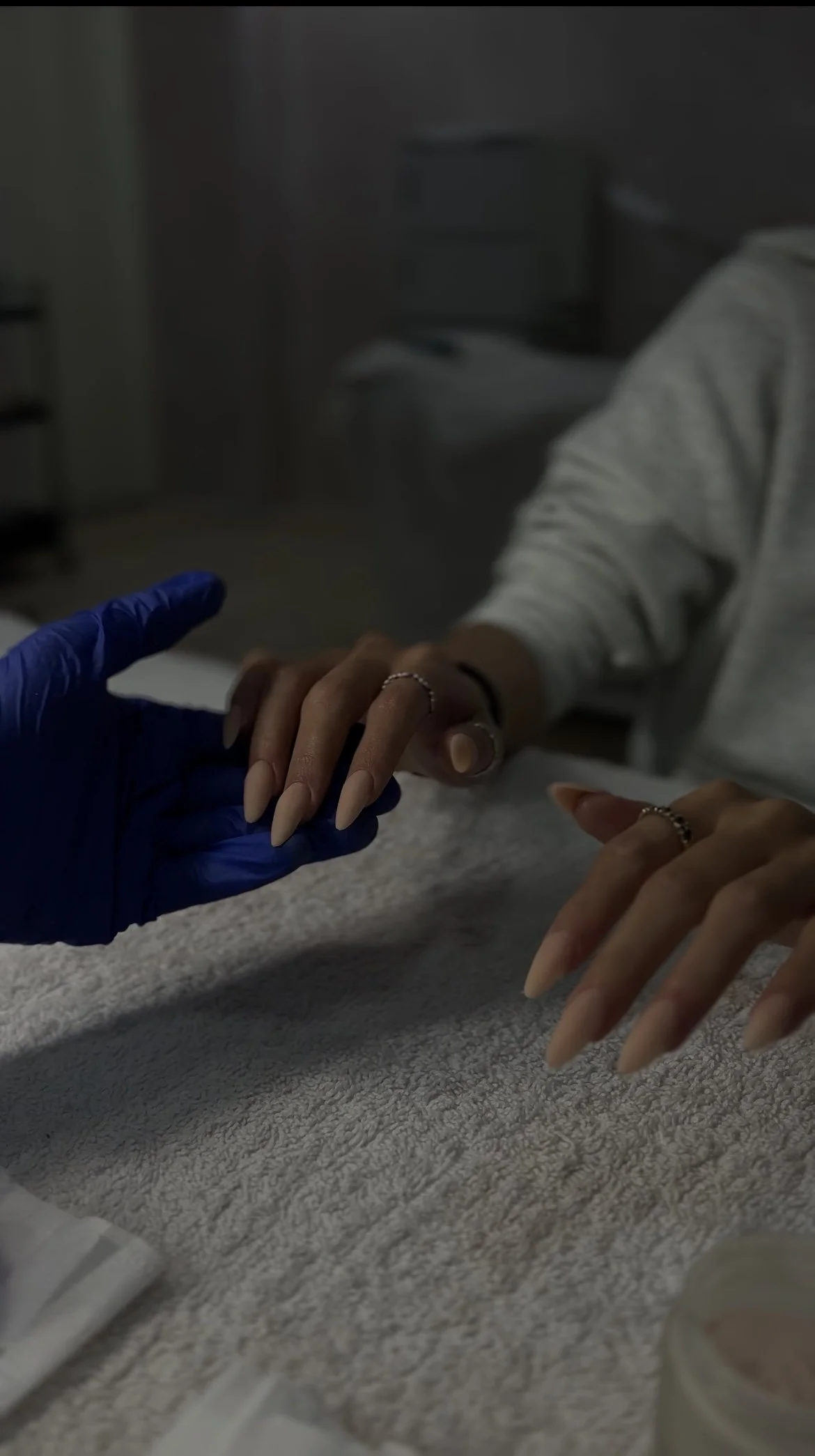 Person receiving a manicure from a nail technician wearing blue gloves, with hands resting on a textured towel.