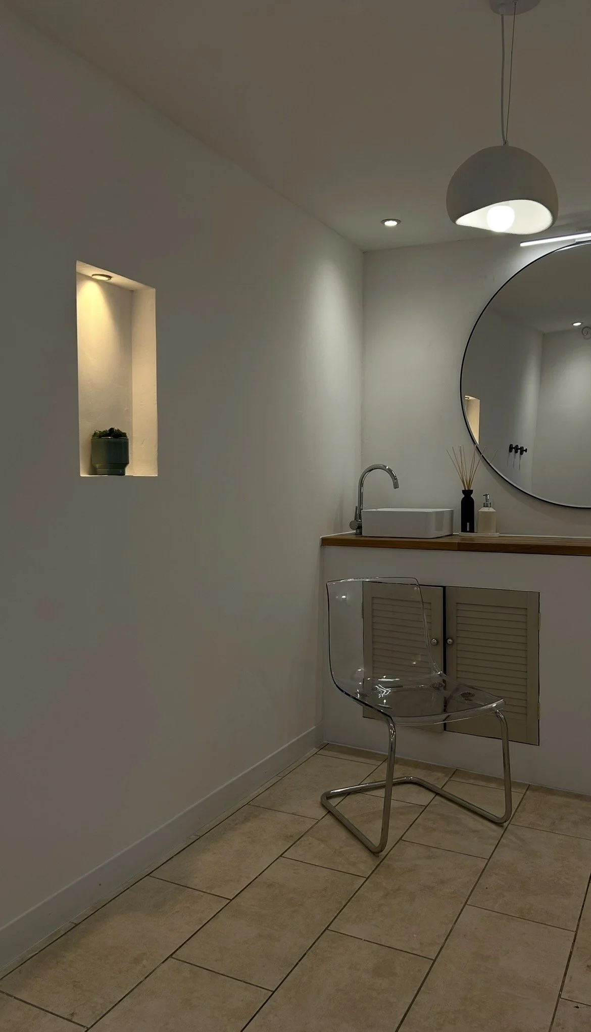 A minimalist bathroom with white walls, a large round mirror, a small countertop sink, a transparent chair, and a decorative shelf with a potted plant. There is a pendant light overhead and recessed lighting.
