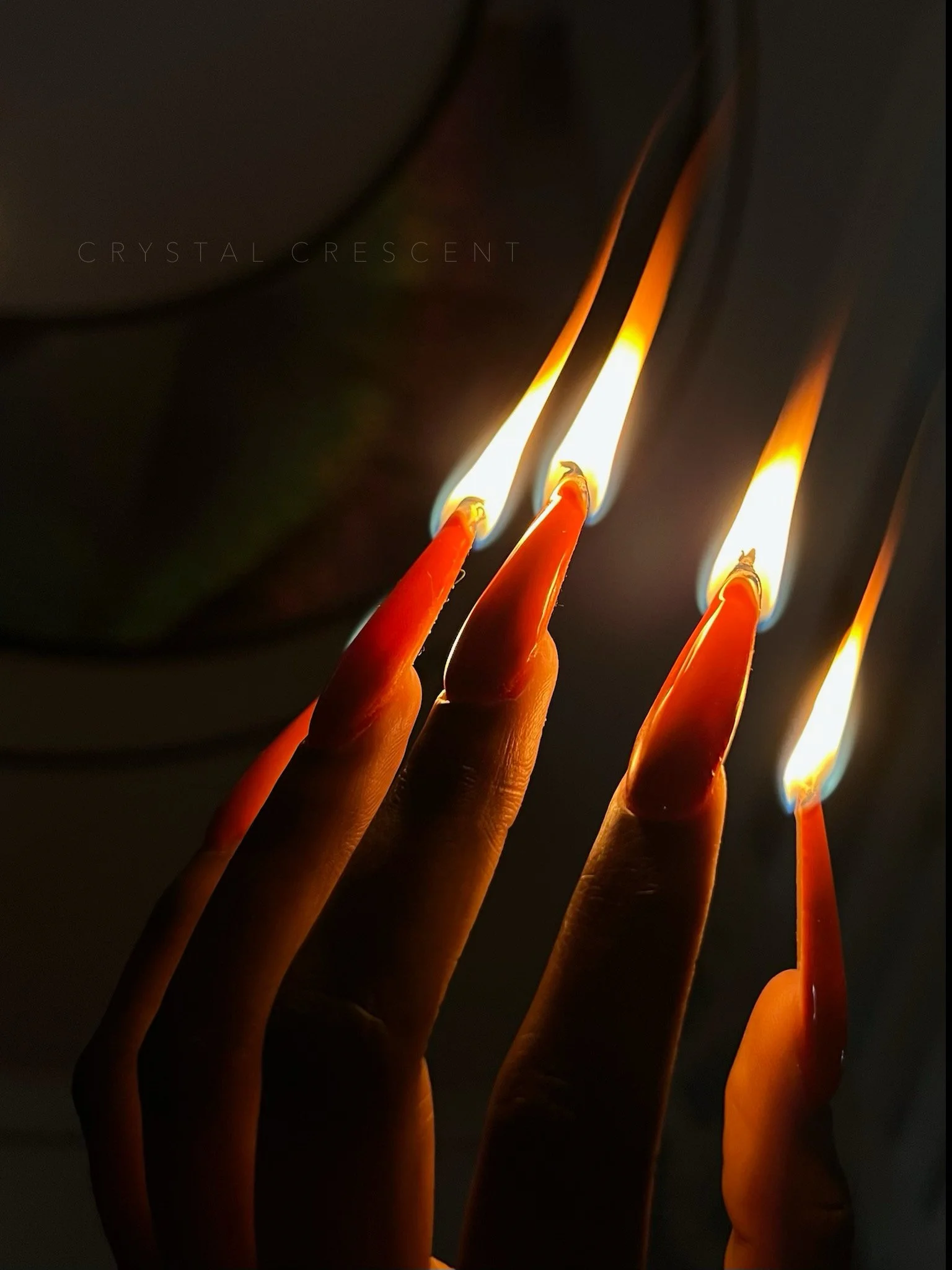 Close-up of four fingers with long red painted nails holding matchsticks with lit flames, dark background.