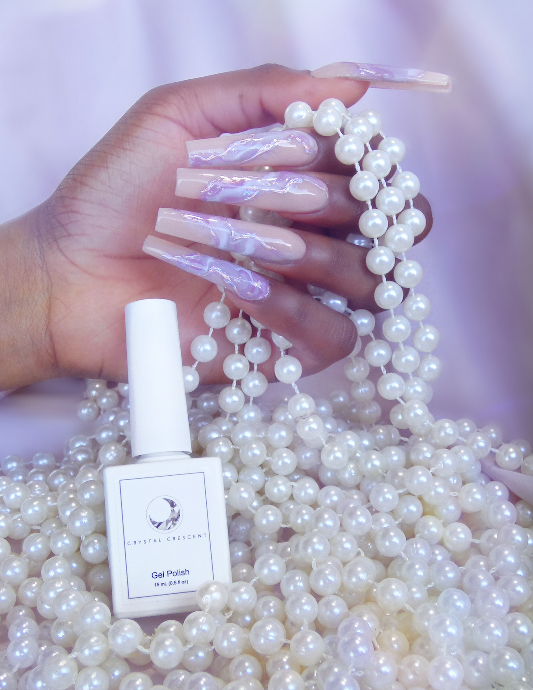 Hand holding long, square-shaped, iridescent nails with pearl necklaces, with a bottle of gel nail polish in the foreground. The background is filled with pearl necklaces.