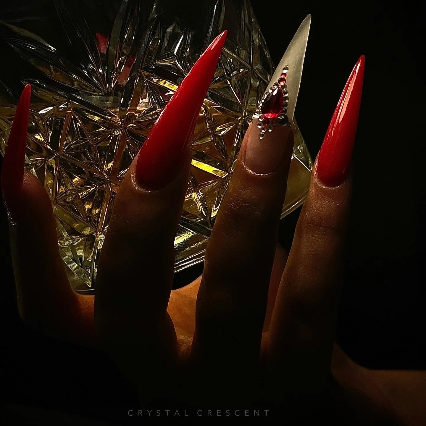 Close-up of a hand with long, sharp red nails holding a faceted crystal glass. The hand has a decorative ring with a red gem and metallic beads. The background is dark.