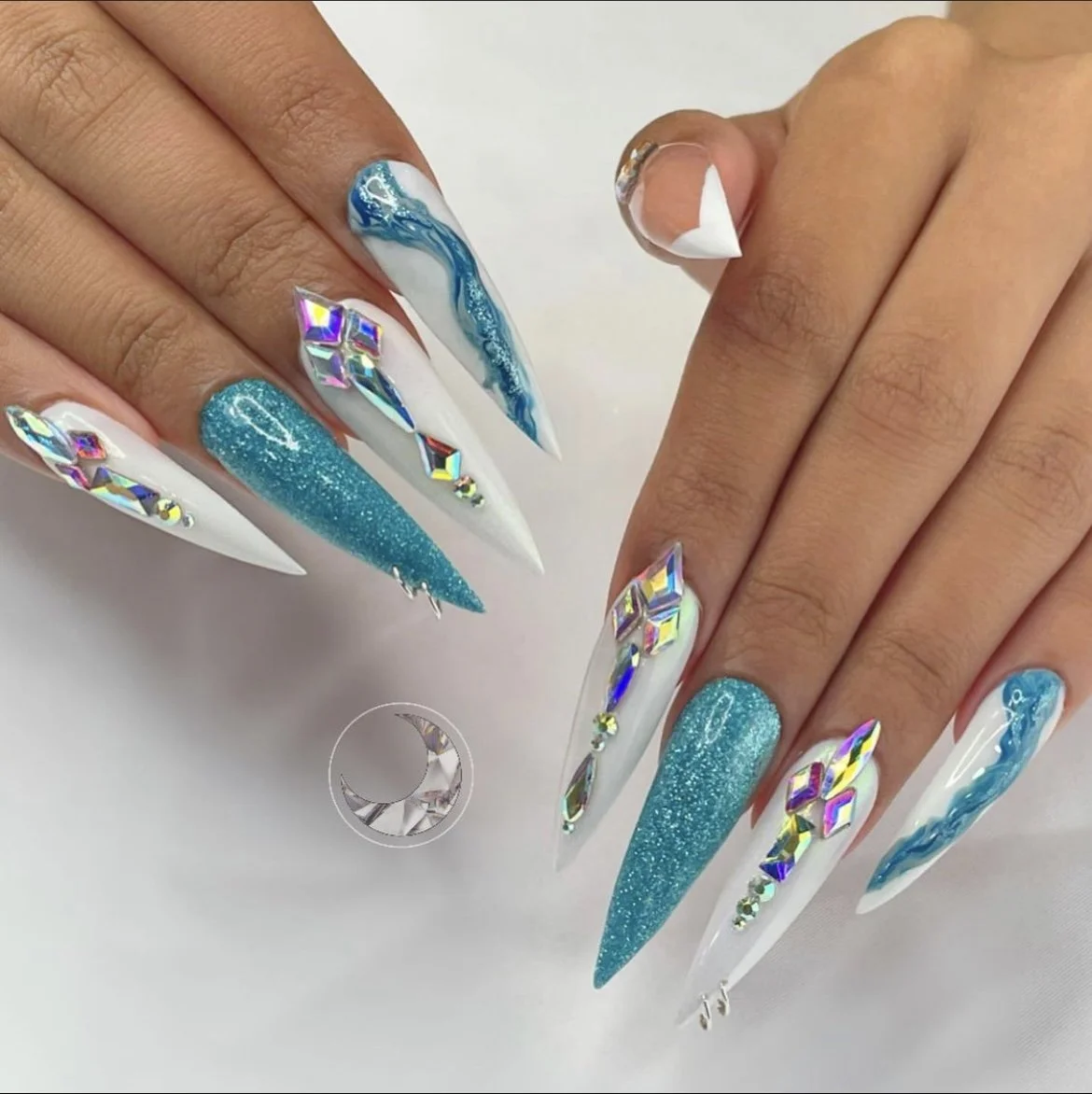 Close-up of a hand with long, pointed acrylic nails in a blue and white marble design, accented with holographic rhinestones and glitter, on a white background.