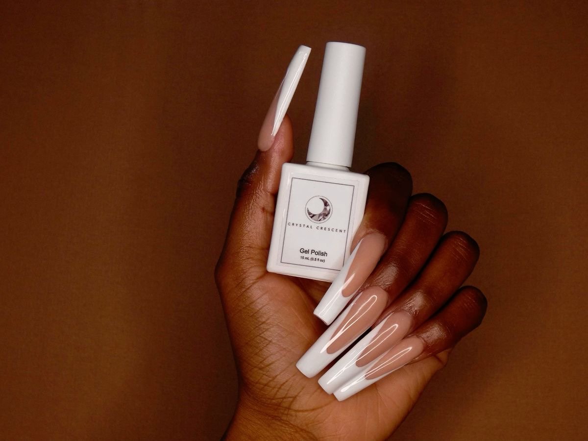 A hand holding a bottle of Crystal Crescent gel nail polish with long, nude-colored, glossy, manicured nails.