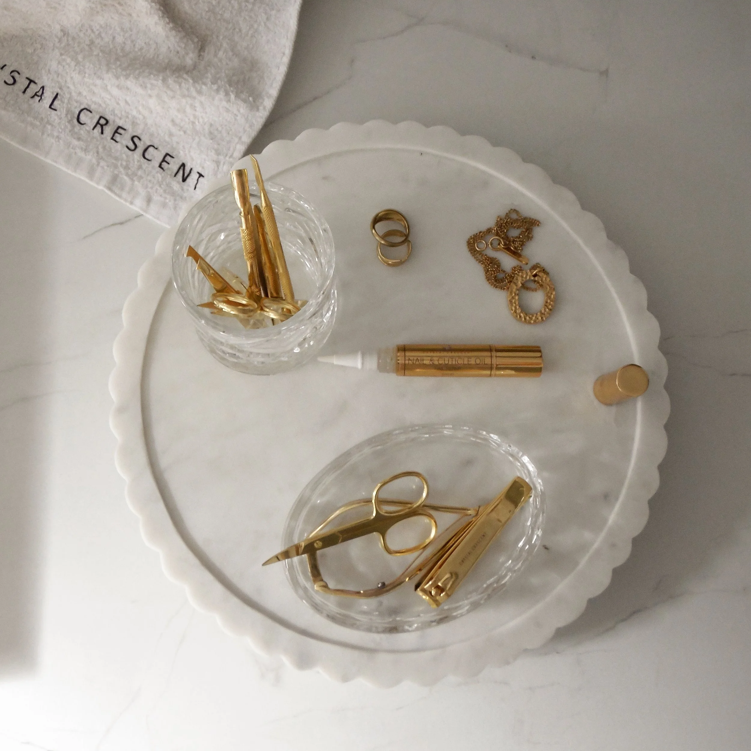 Gold manicure tools, rings, and jewelry on a white scalloped tray, with a towel partially visible in the top left corner.