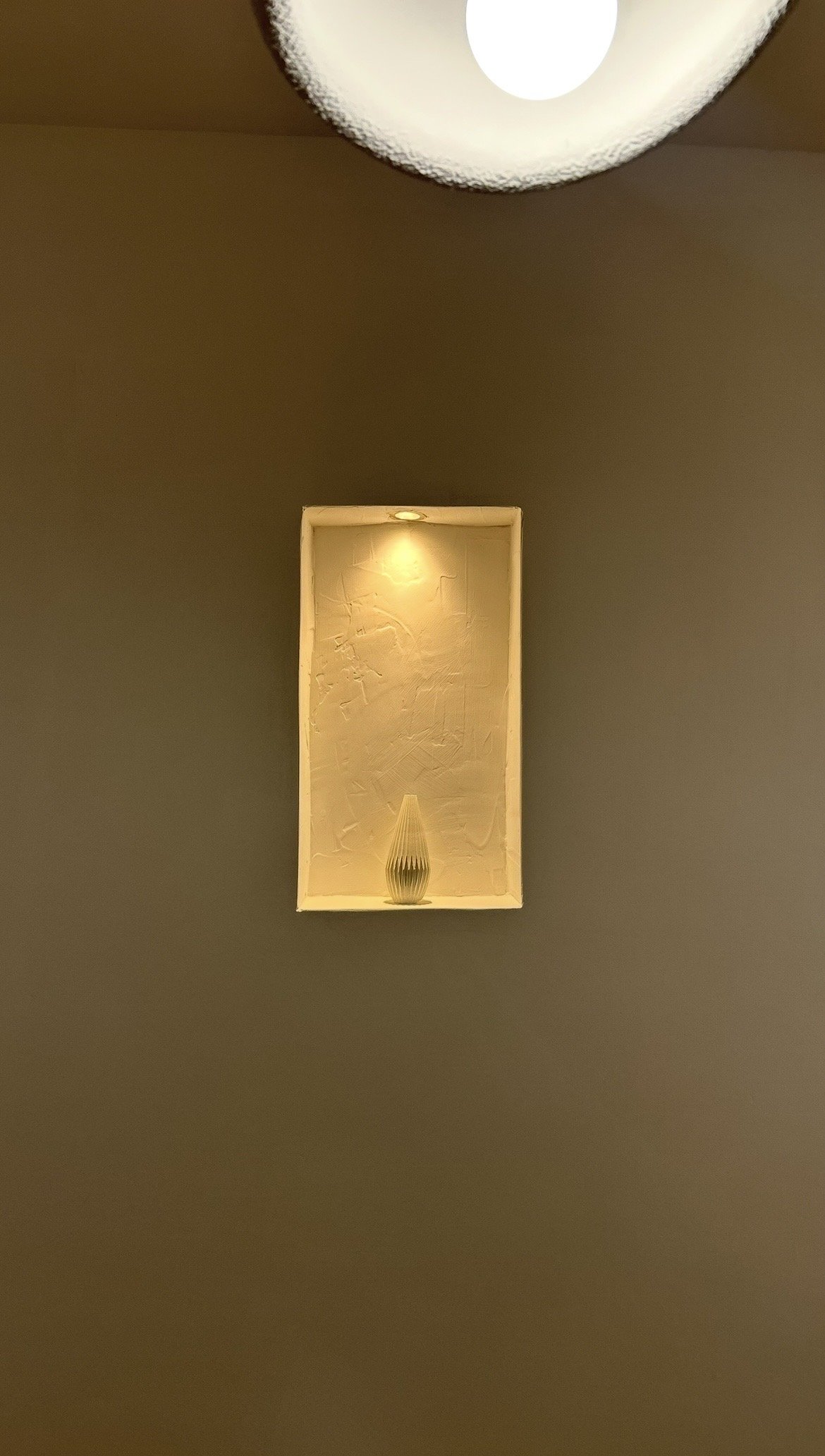 A decorative shelf with a small vase, mounted on a wall with textured beige background, illuminated by a spotlight.