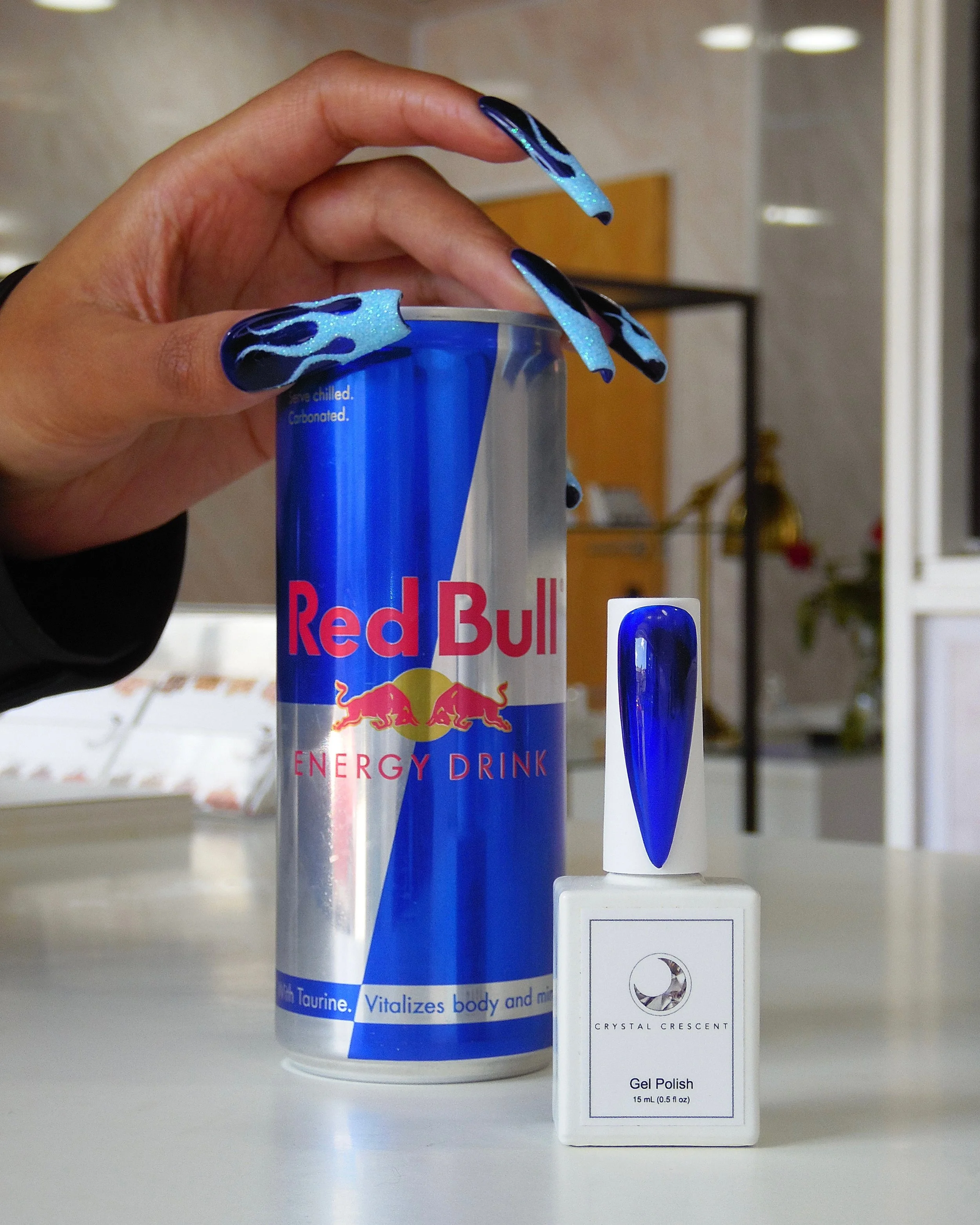 A person's hand with elaborate blue and black flame nail art holds a can of Red Bull energy drink. In front of the can is a bottle of crystal crescent gel polish with blue color, placed on a white surface in a room with shelves and plants in the back