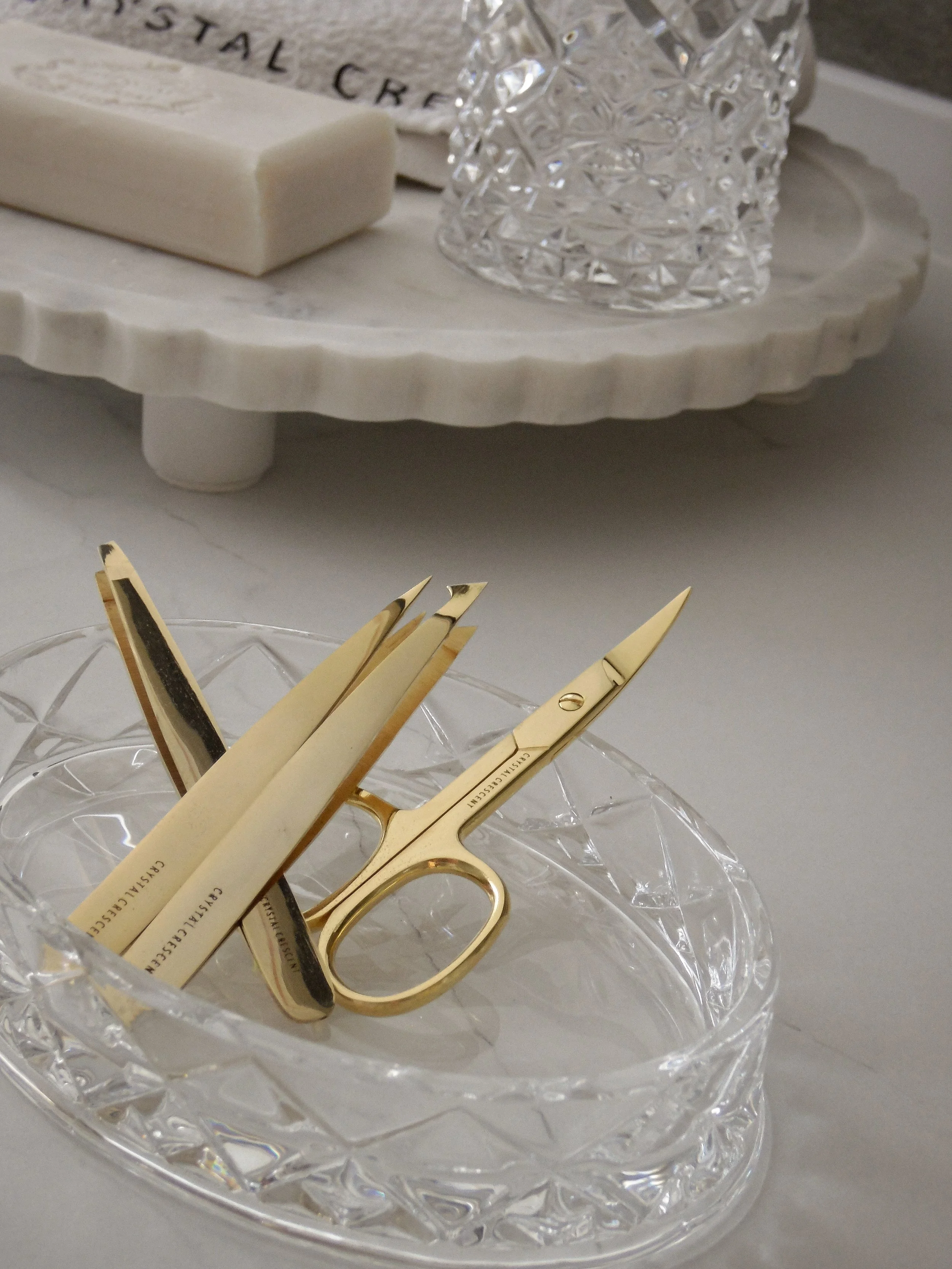 Gold manicure scissors and nail files in a glass dish on a light-colored surface, with a marble tray and crystal glass in the background.
