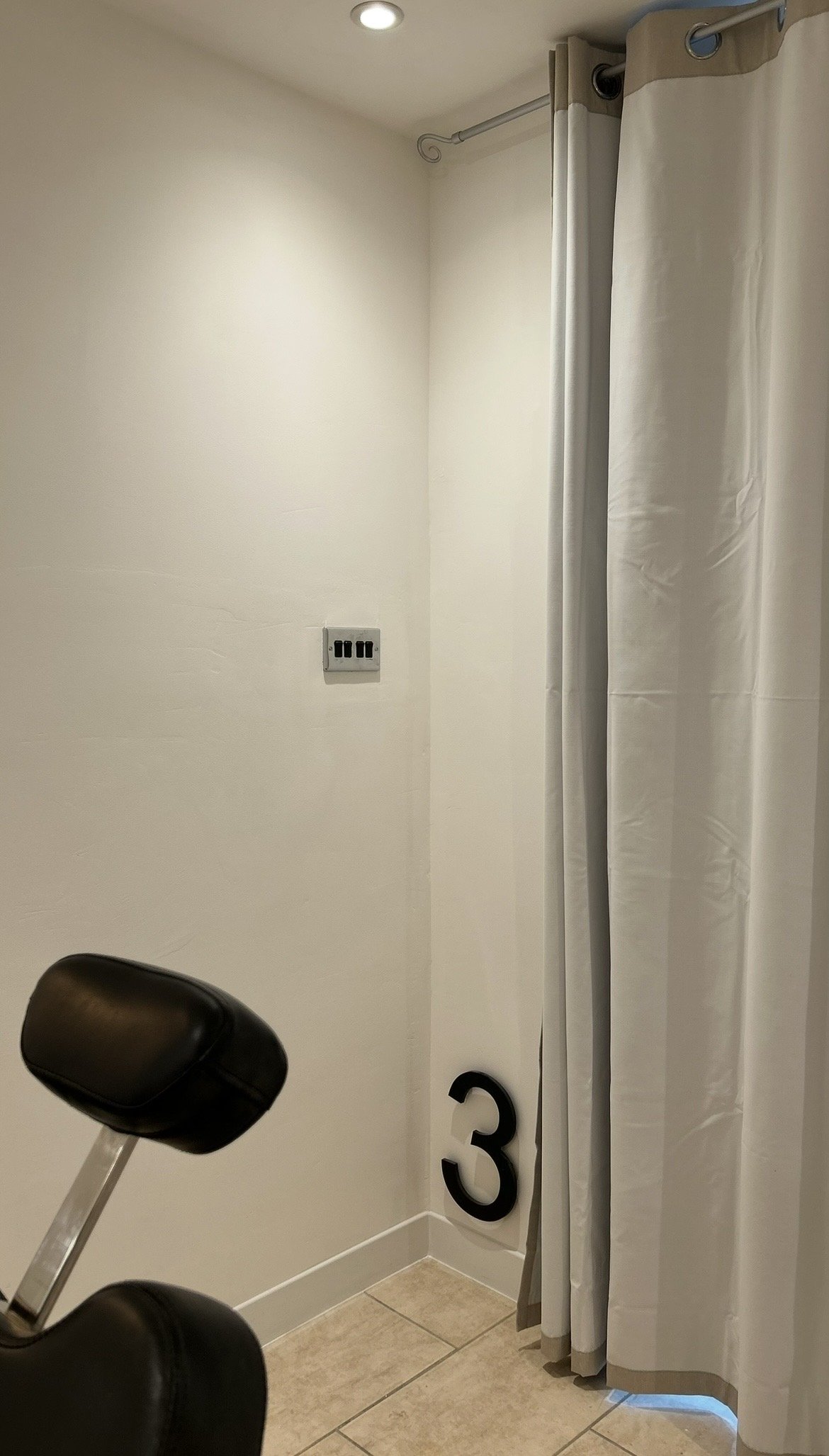 A corner of a room with a beige tiled floor, white walls, a black chair with a metal frame, and a white curtain hanging from a curtain rod. There is a black number 3 decal on the wall. An electrical outlet with three switches is visible on the wall, 