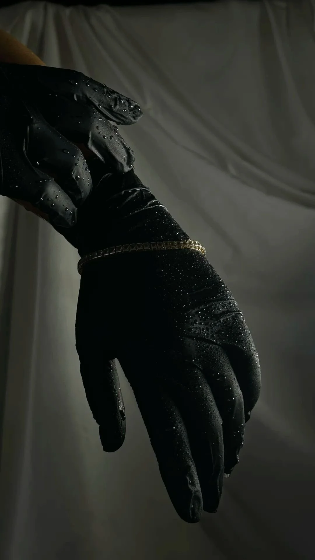 A hand wearing a black glove with water droplets on it, holding a gold chain.