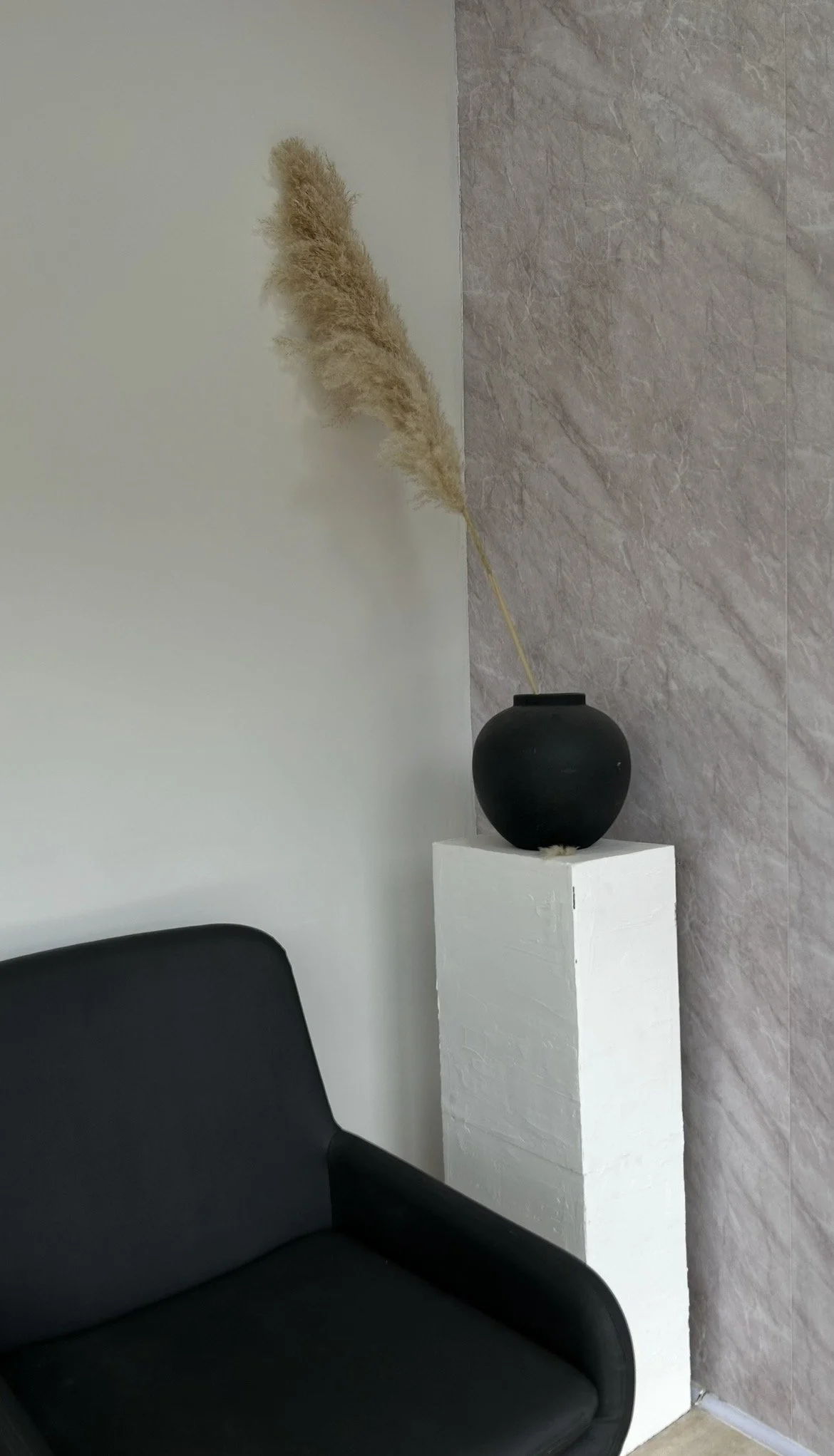 A black sofa in front of a white wall, with a white pedestal holding a black vase containing beige pampas grass, against a textured grey wall.